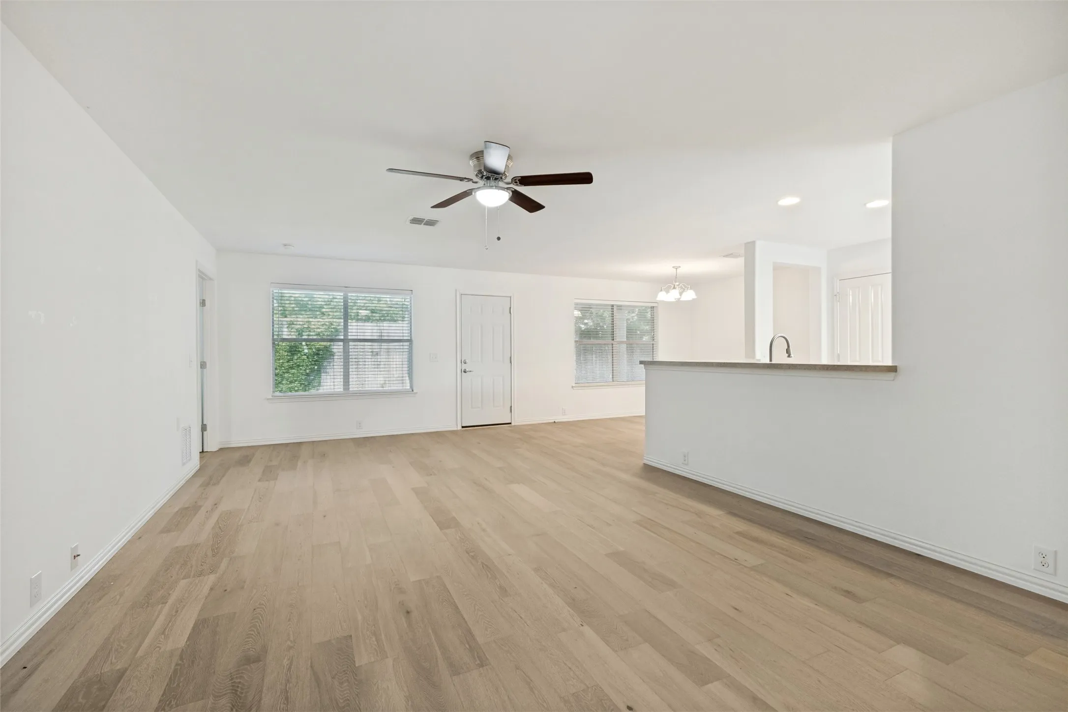Expansive living area featuring light wood-finish flooring, a ceiling fan, and white painted walls overlooking the dining area and kitchen.