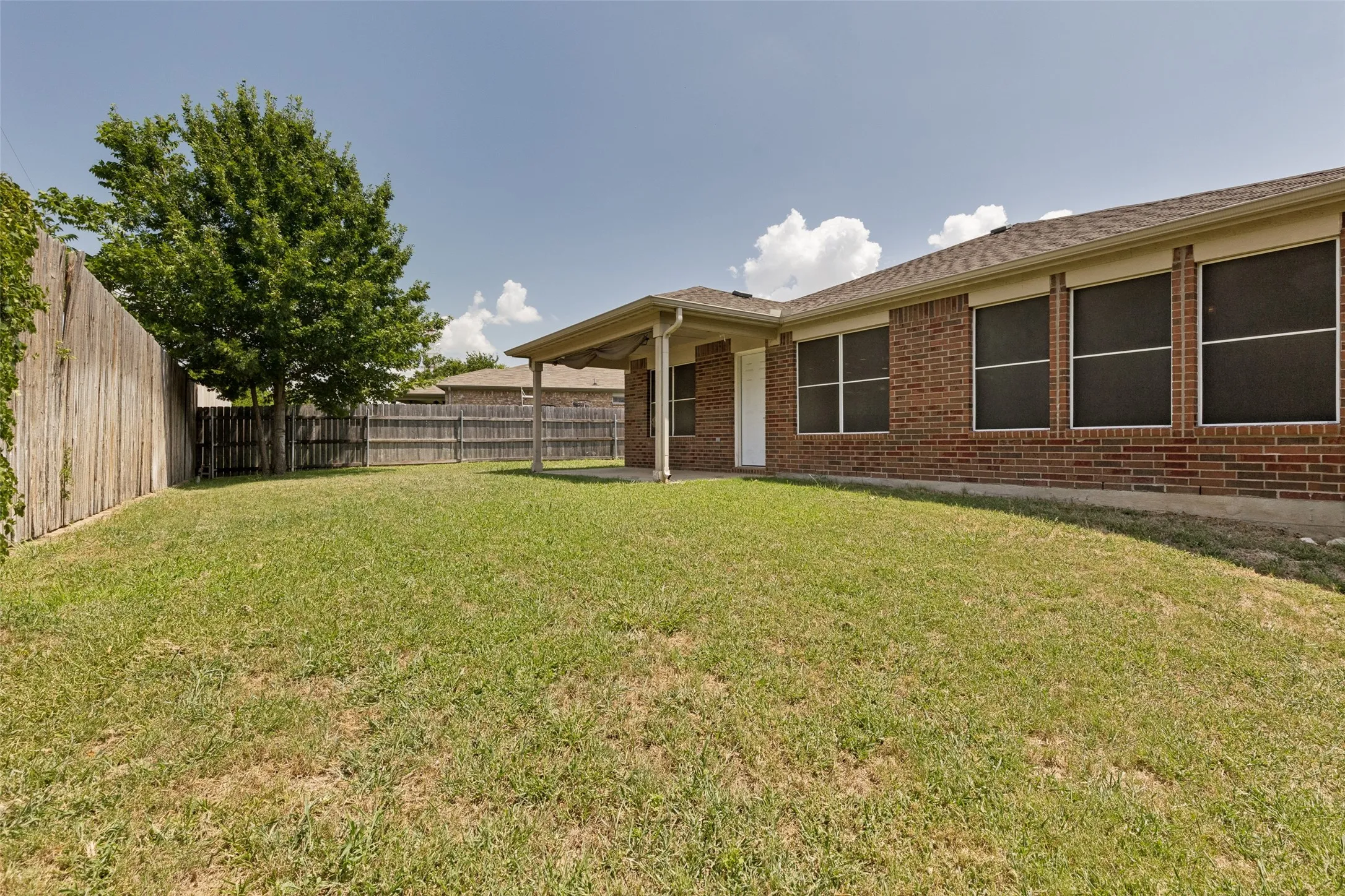 Expansive backyard featuring a covered patio, mature tree, full privacy fencing, and a brick exterior with multiple windows
