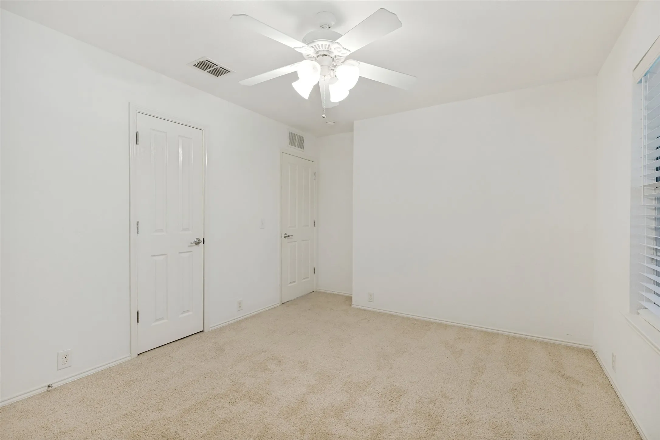 Spacious room featuring neutral carpeting, white walls, a multi-pane window with horizontal blinds, and a white ceiling fan with integrated lighting