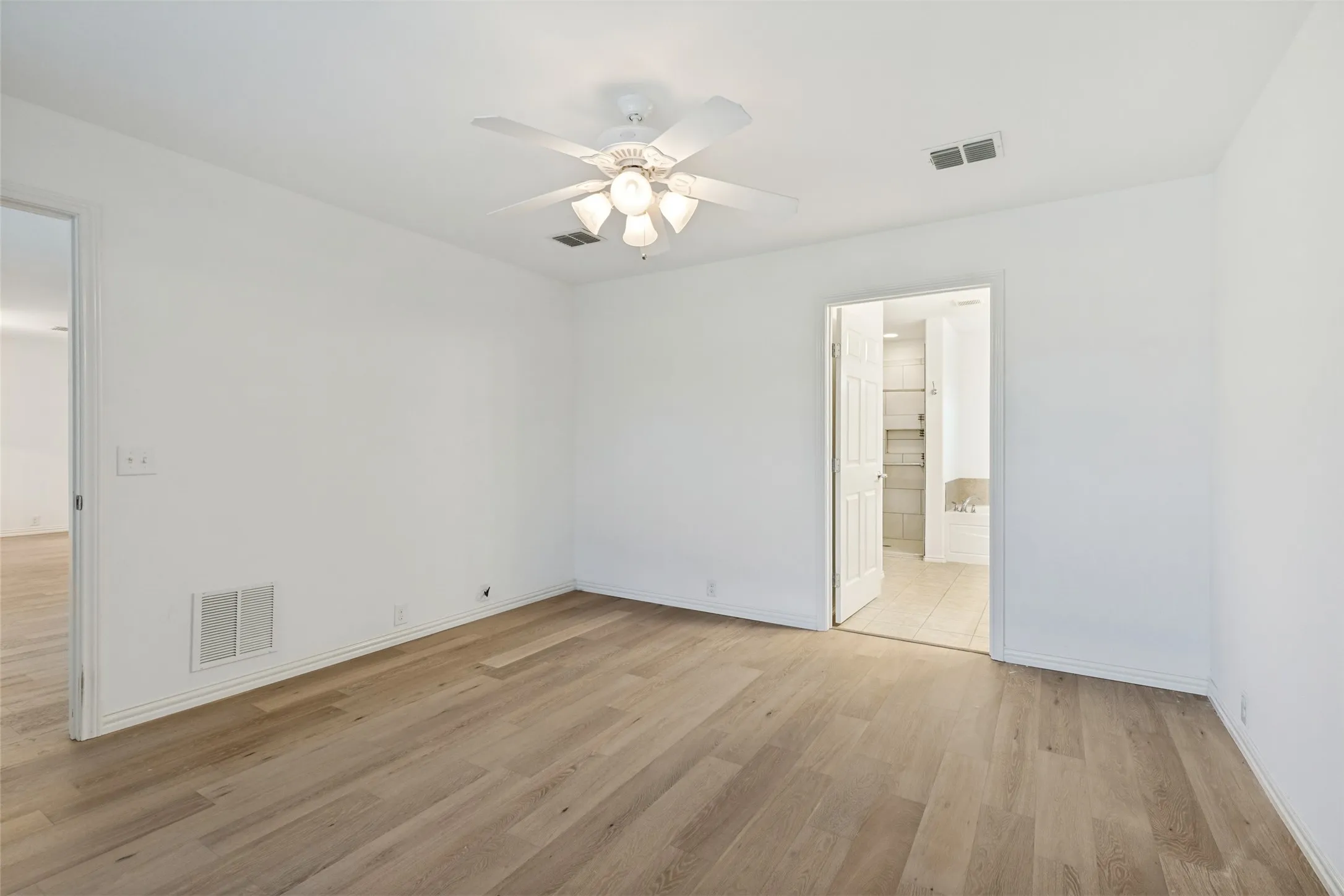 Spacious primary bedroom featuring light wood-finish looring, white walls, and a ceiling fan with integrated lighting