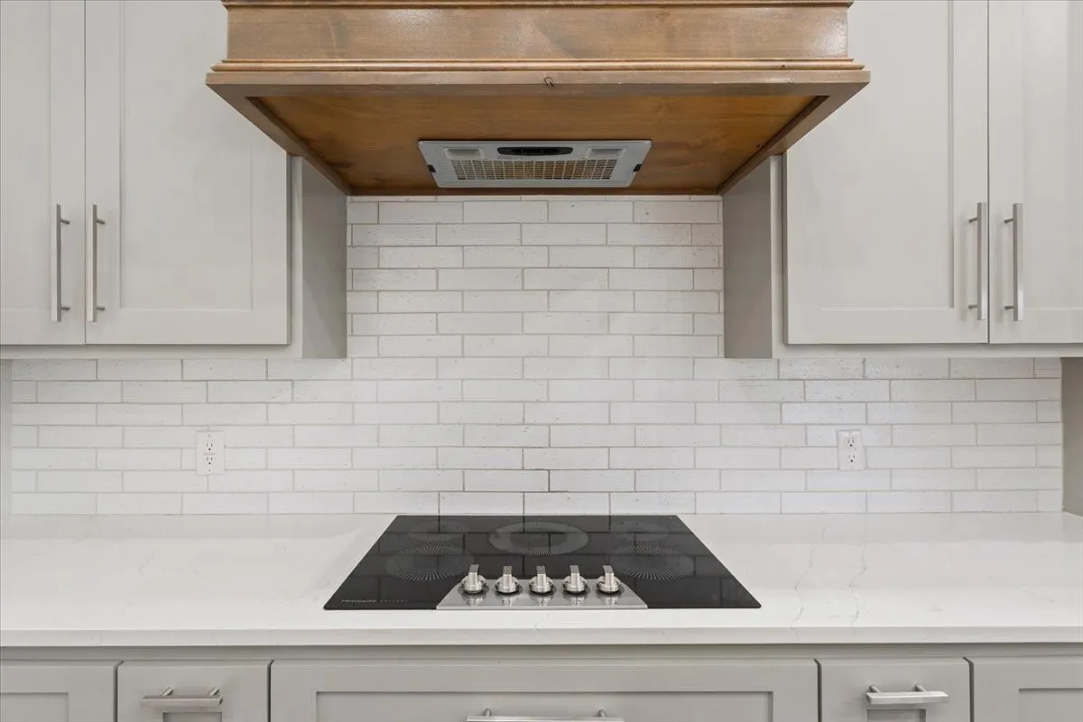 Kitchen featuring a built-in cooktop, light gray cabinetry with brushed nickel pulls, white subway tile backsplash, light-toned countertops, and a wood-finish range hood