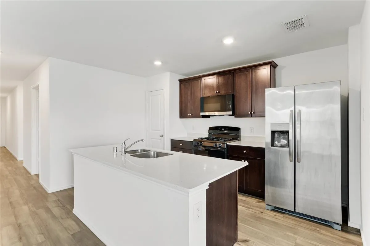 Modern kitchen featuring a spacious white quartz island with waterfall edge, stainless steel side-by-side refrigerator with ice and water dispenser, dark wood cabinetry with contrasting white upper and lower countertops, gas cooktop with range hood, and wood-finish flooring throughout the open layout