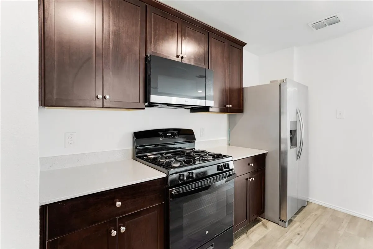 Modern kitchen featuring rich wood-finish cabinetry with integrated hardware, stainless steel gas range with multiple burners and convection oven, built-in microwave, white quartz countertops, wood-finish flooring, and stainless steel refrigerator with ice and water dispenser