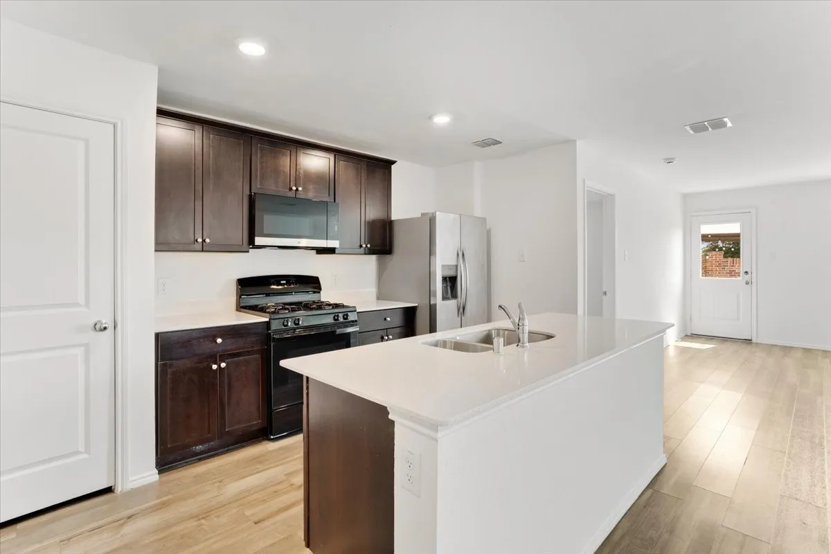 Kitchen featuring a white countertop island with an undermount dual-basin sink, dark wood-finish cabinetry, stainless steel refrigerator, and black gas range