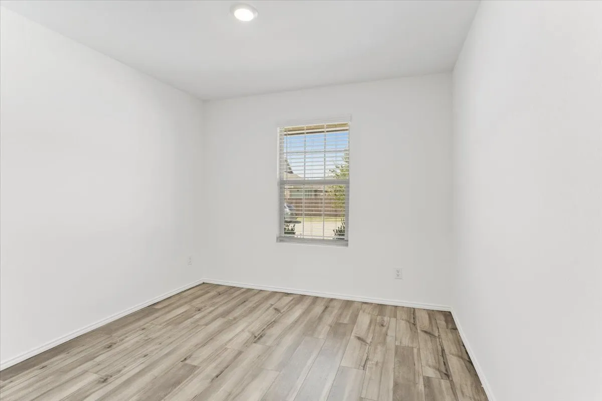 Bright bedroom featuring wood-finish flooring, white walls, and a single-hung window with horizontal blinds allowing natural light throughout the space