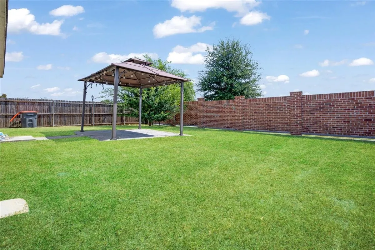 Expansive backyard oasis featuring a professionally manicured lawn, freestanding pergola with integrated shade structure, mature landscaping with established trees, brick perimeter fencing for privacy, and open grounds ideal for outdoor entertaining and recreation