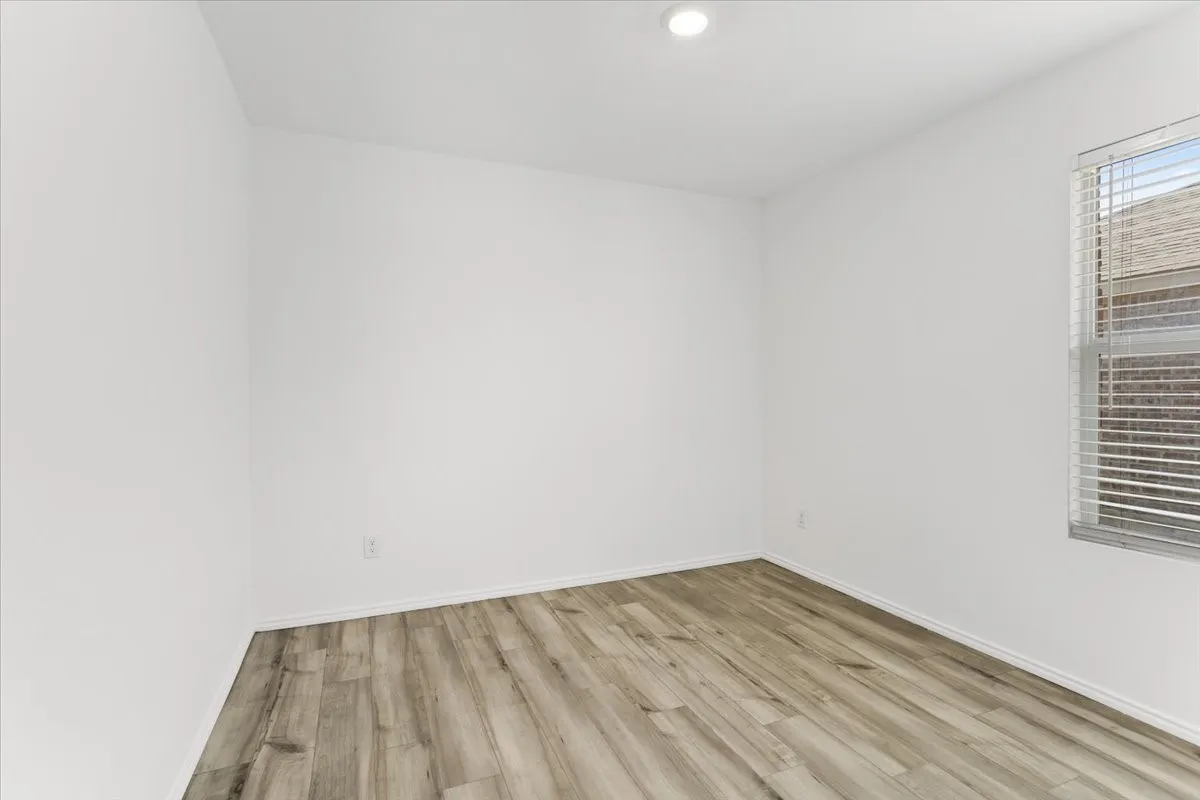 Spacious bedroom showcasing warm wood-finish flooring, clean white walls, and abundant natural light from a large window with horizontal blinds