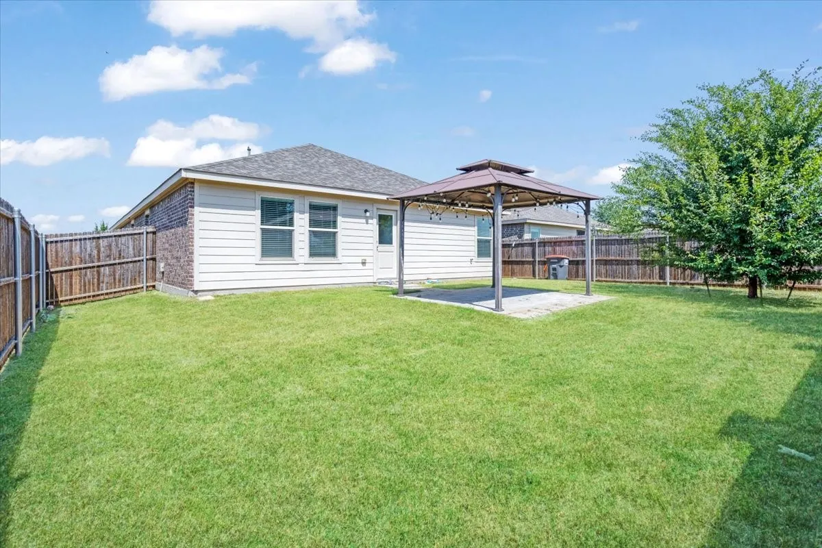 Spacious backyard featuring a manicured lawn with mature trees, covered patio structure, and wooden fencing