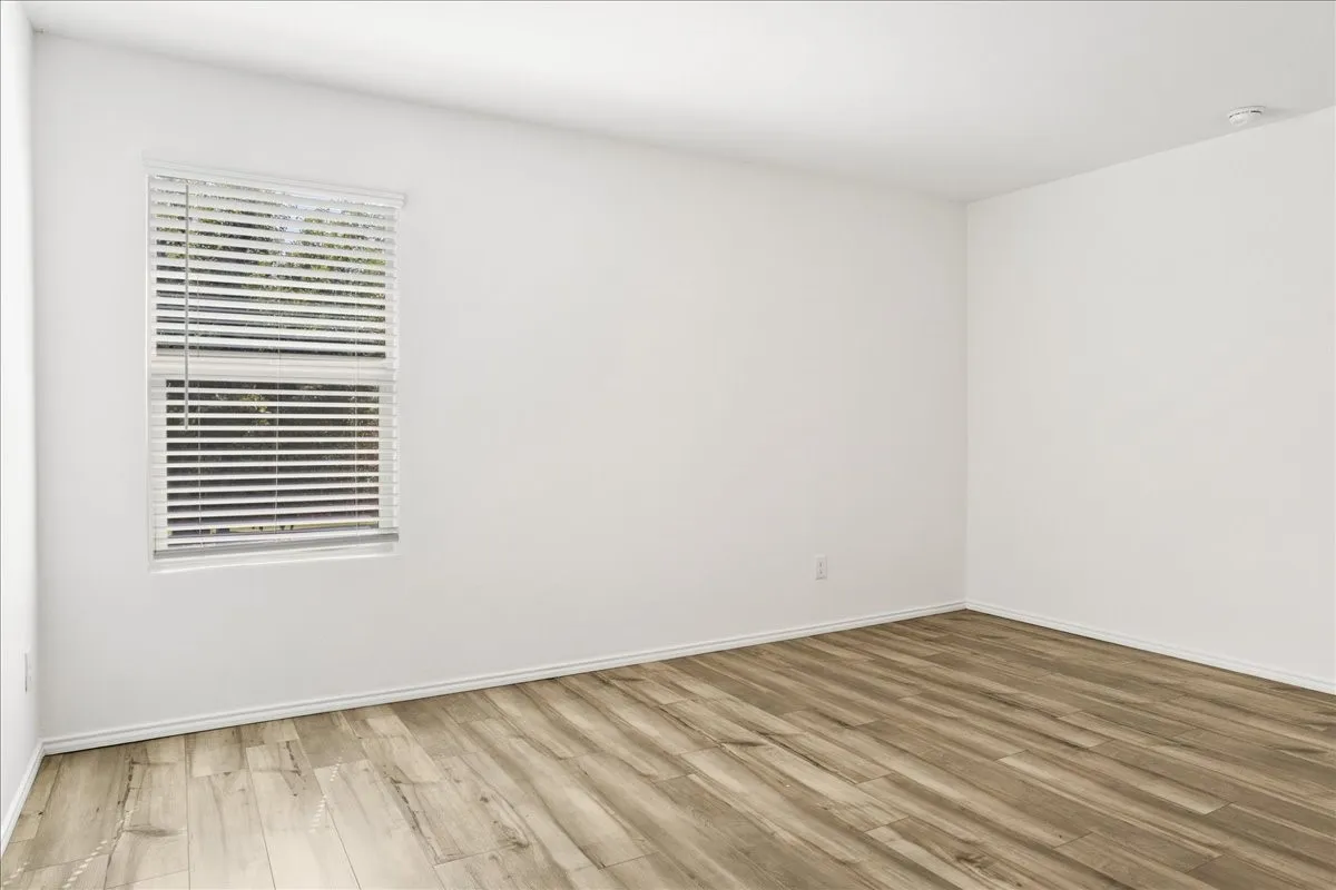 Bright corner bedroom showcasing pristine white walls, a large double-hung window with plantation shutters providing natural light and ventilation, and newly installed wood-finish plank flooring throughout the space