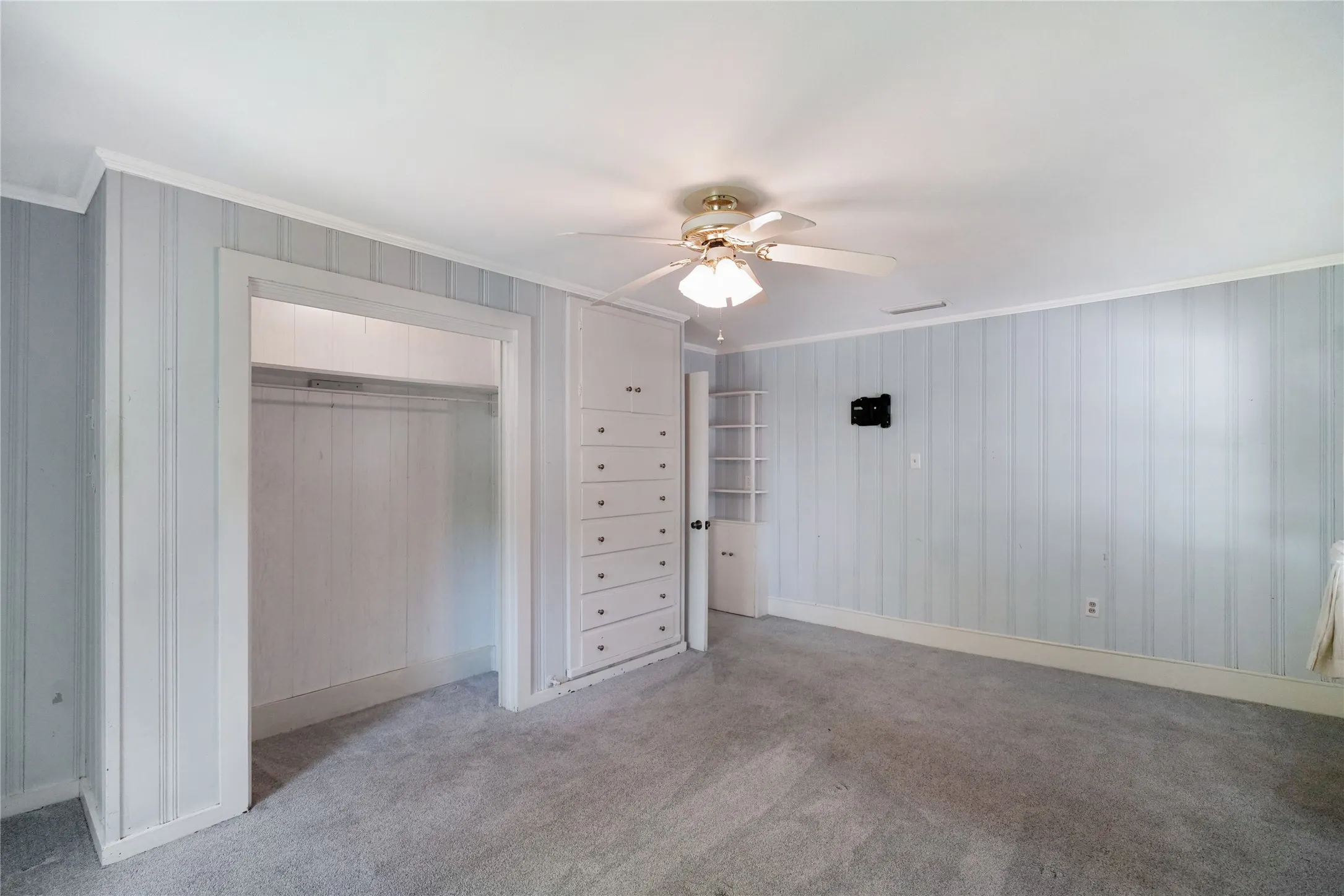 Room featuring textured wall paneling, ceiling fan with light fixture, built-in dresser, built-in shelving, and carpet flooring