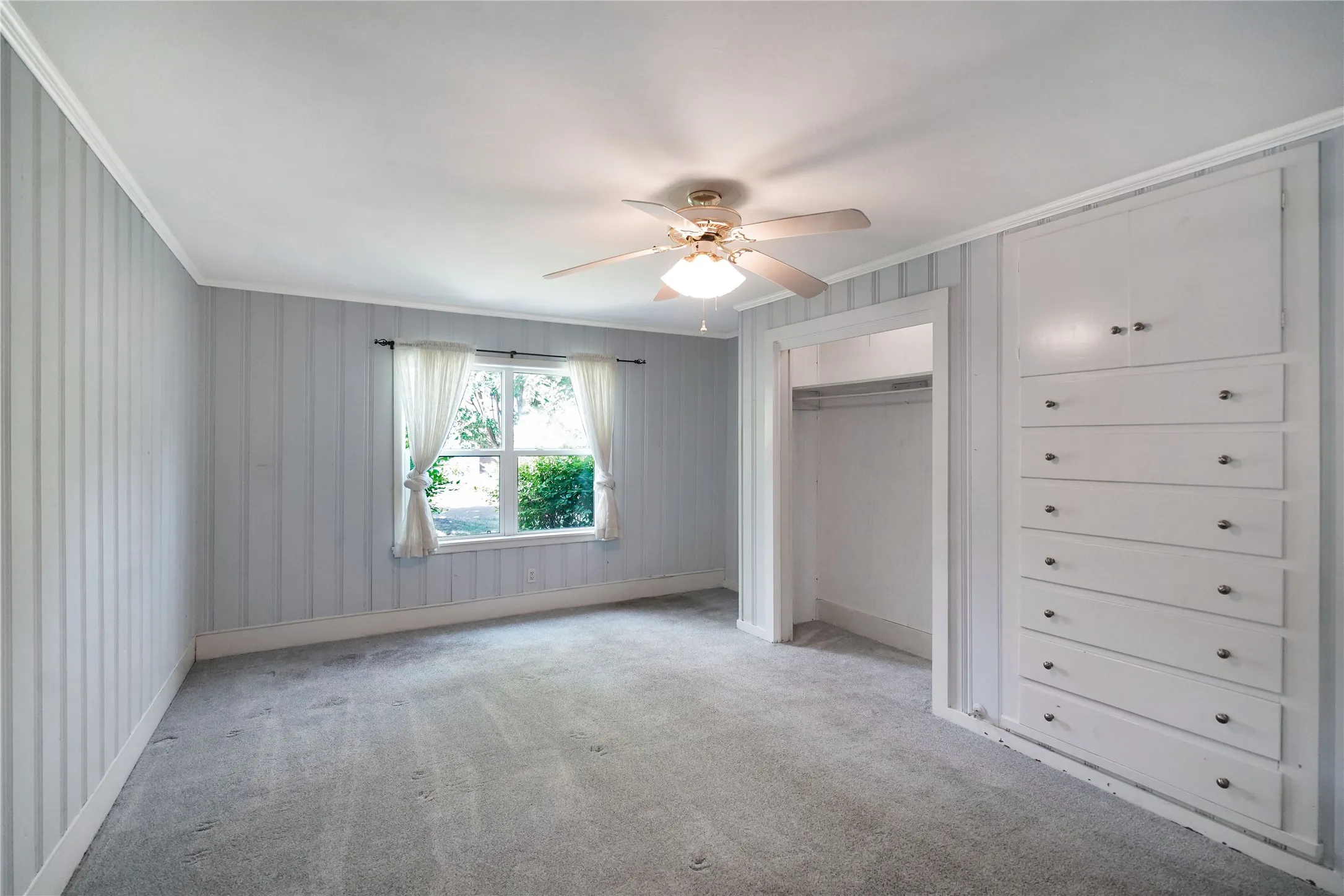 Room featuring light blue vertical paneling, grey carpet, a ceiling fan with integrated lighting, and a built-in storage unit with drawers and cabinets