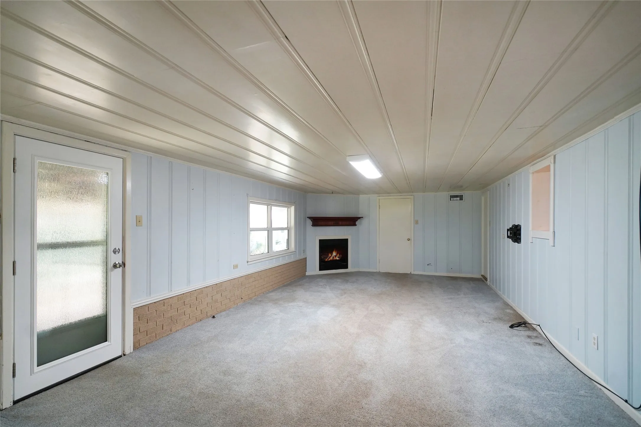 Spacious interior featuring a corner fireplace with a dark wood mantel, light blue paneling, a brick base wall, carpet flooring, and a glass-paned exterior door