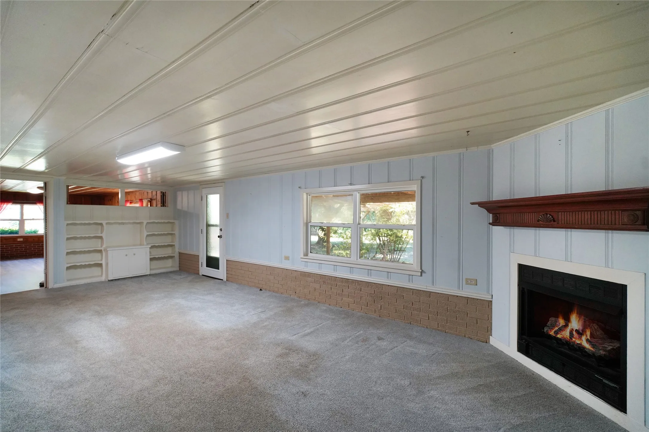 Spacious interior room featuring light-colored carpeting, a fireplace with a wood mantle, and light blue vertical paneling