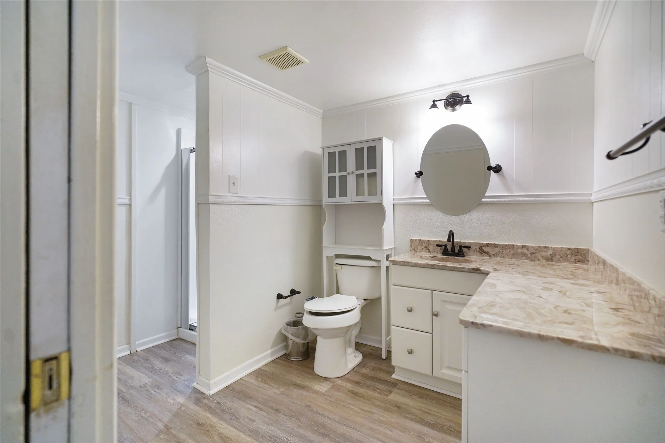 Bathroom featuring a wood-finish floor, wainscoting, an oval mirror, a vanity with under-sink storage, and a shower enclosure