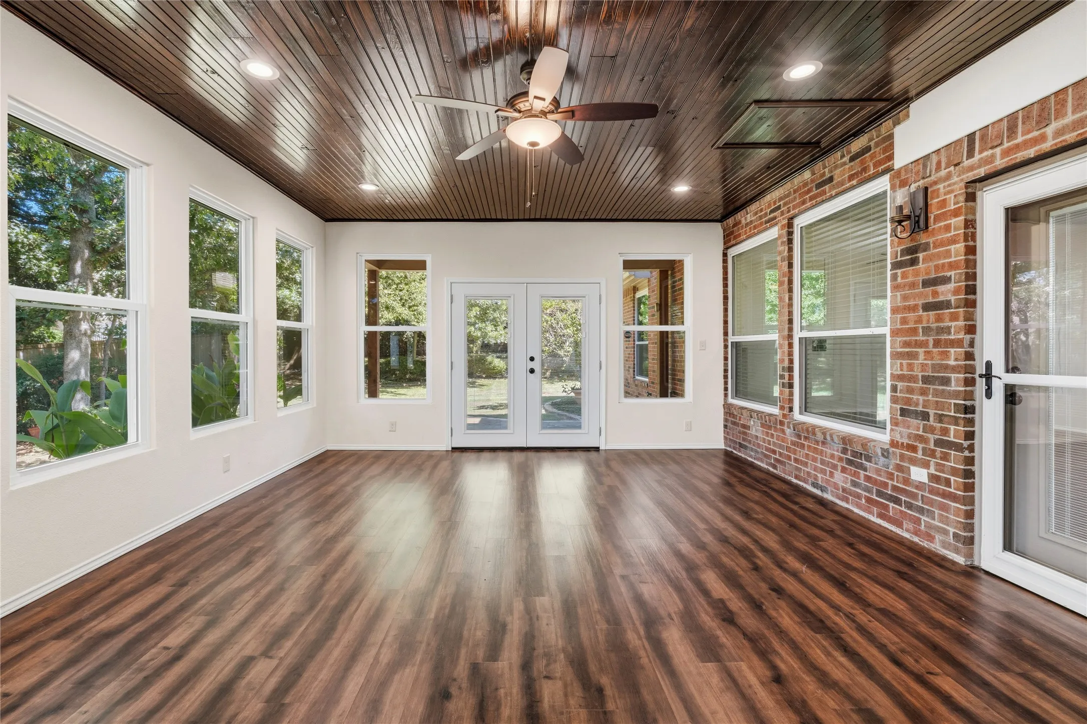 Spacious sunroom featuring wood-finish flooring, a wood-paneled ceiling with recessed lighting and a ceiling fan, a brick accent wall, and multiple windows and glass-paned doors