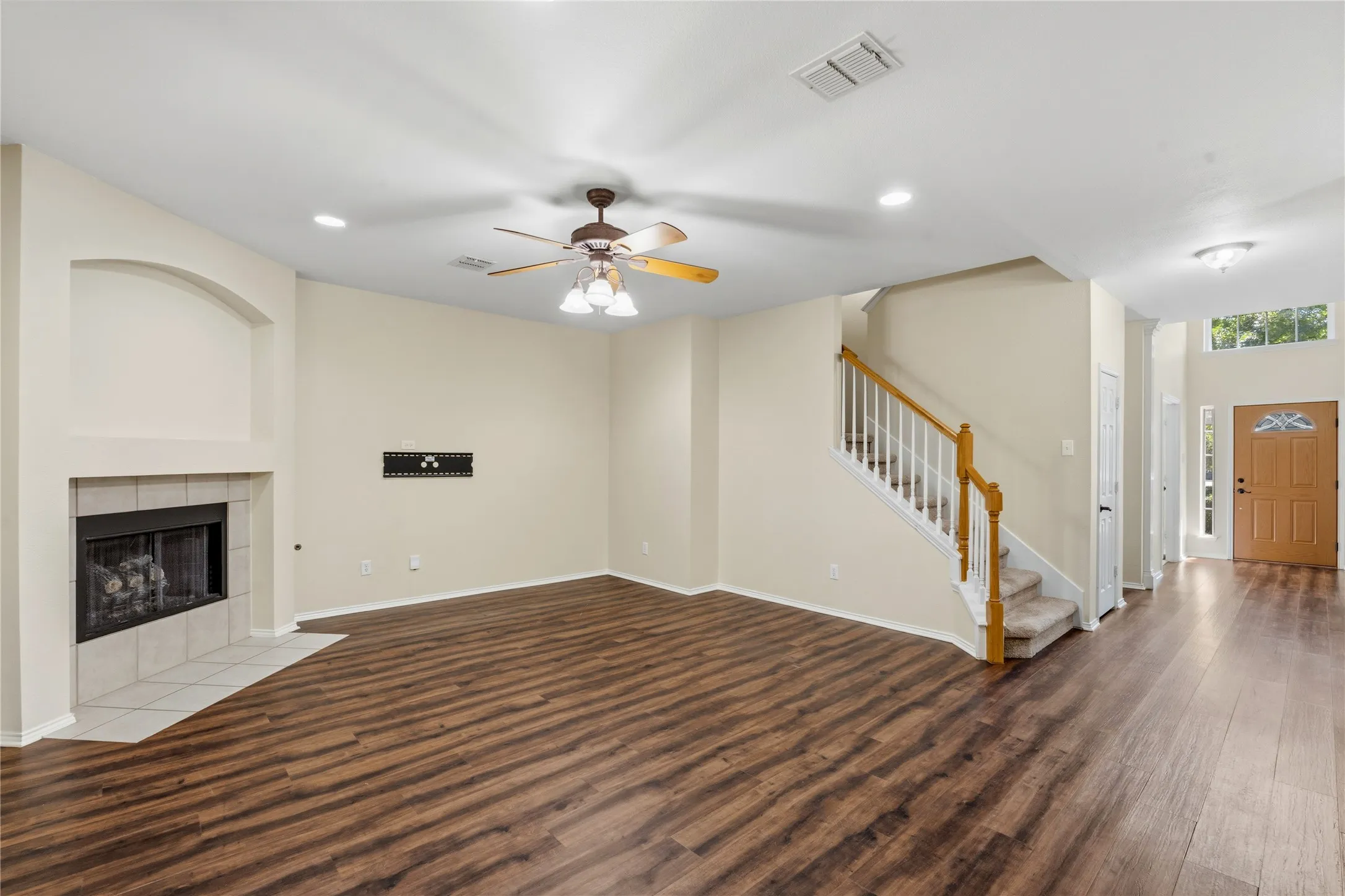 Spacious living area featuring wood-finish flooring, a tiled fireplace with an arched alcove, and a ceiling fan with integrated lighting