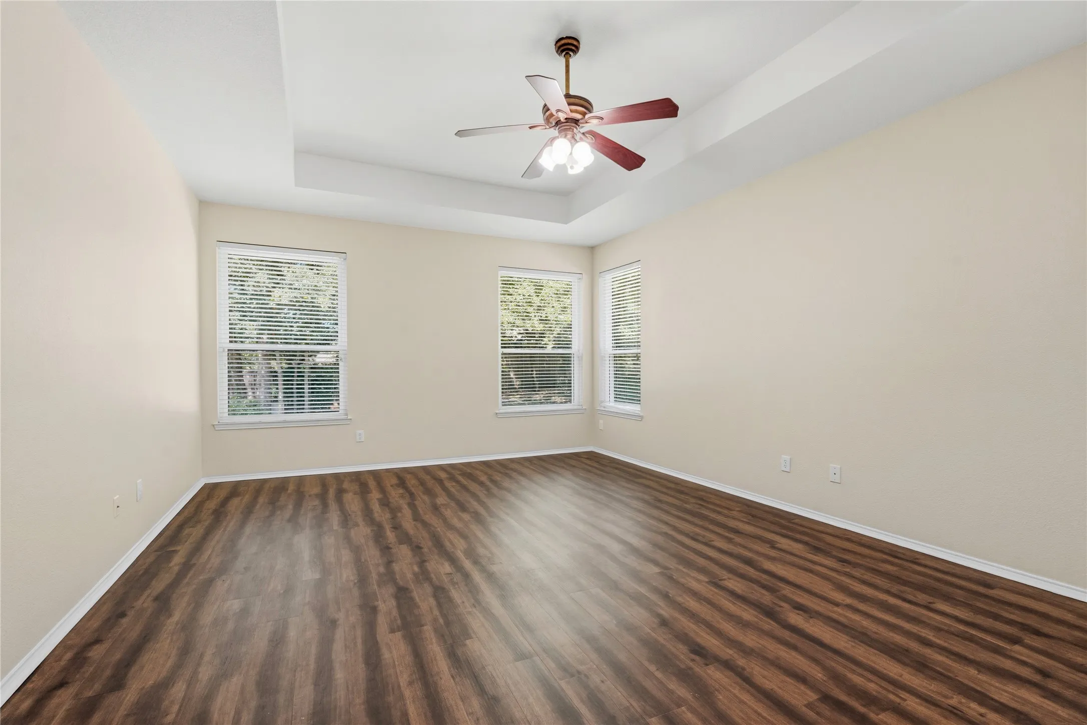 Spacious interior room with wood-finish flooring, three windows with blinds, a tray ceiling, and a ceiling fan with light fixture