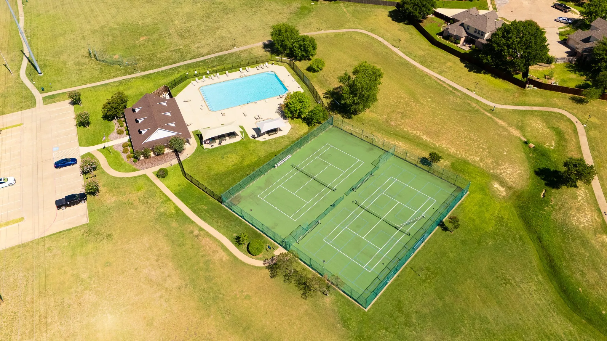 Community recreation area featuring an outdoor swimming pool, two tennis courts, a clubhouse, and paved walking paths surrounded by expansive green spaces