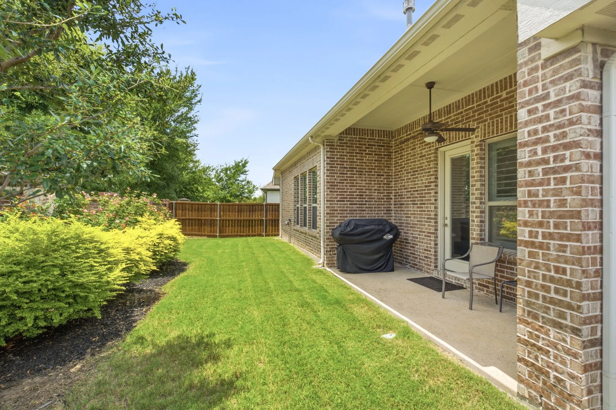 Ceiling fan on covered patio, steel posts in fence on sides of yard, brick wall in back. Lovely landscape beds.