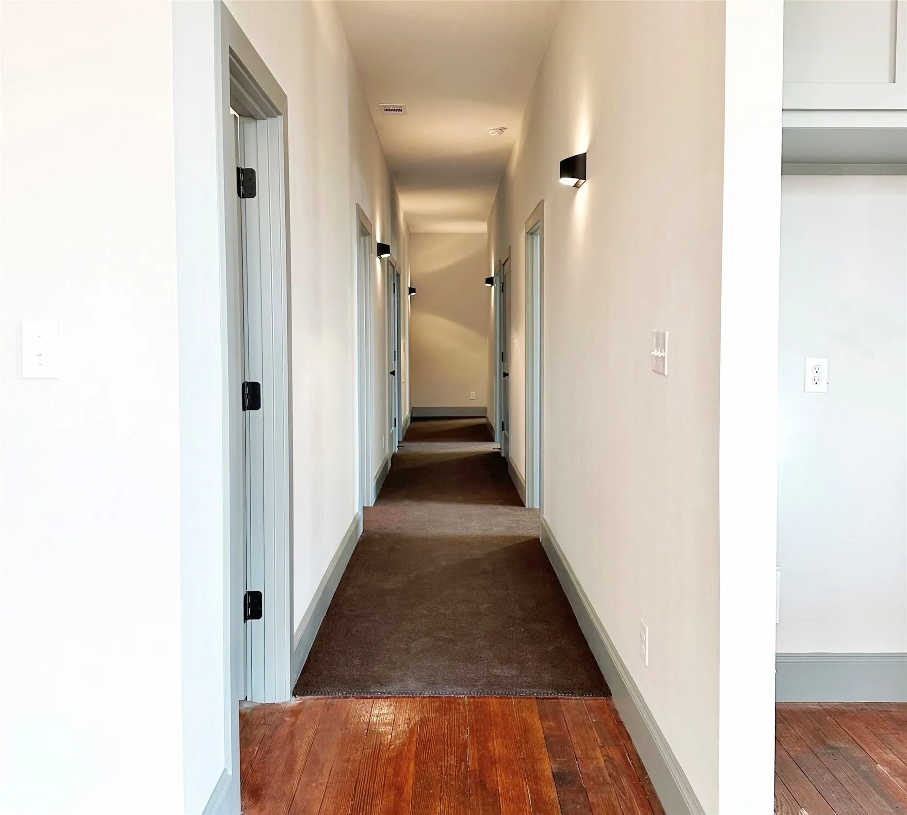 Long hallway featuring wood-finish flooring, light gray walls, and multiple doorways with dark hardware