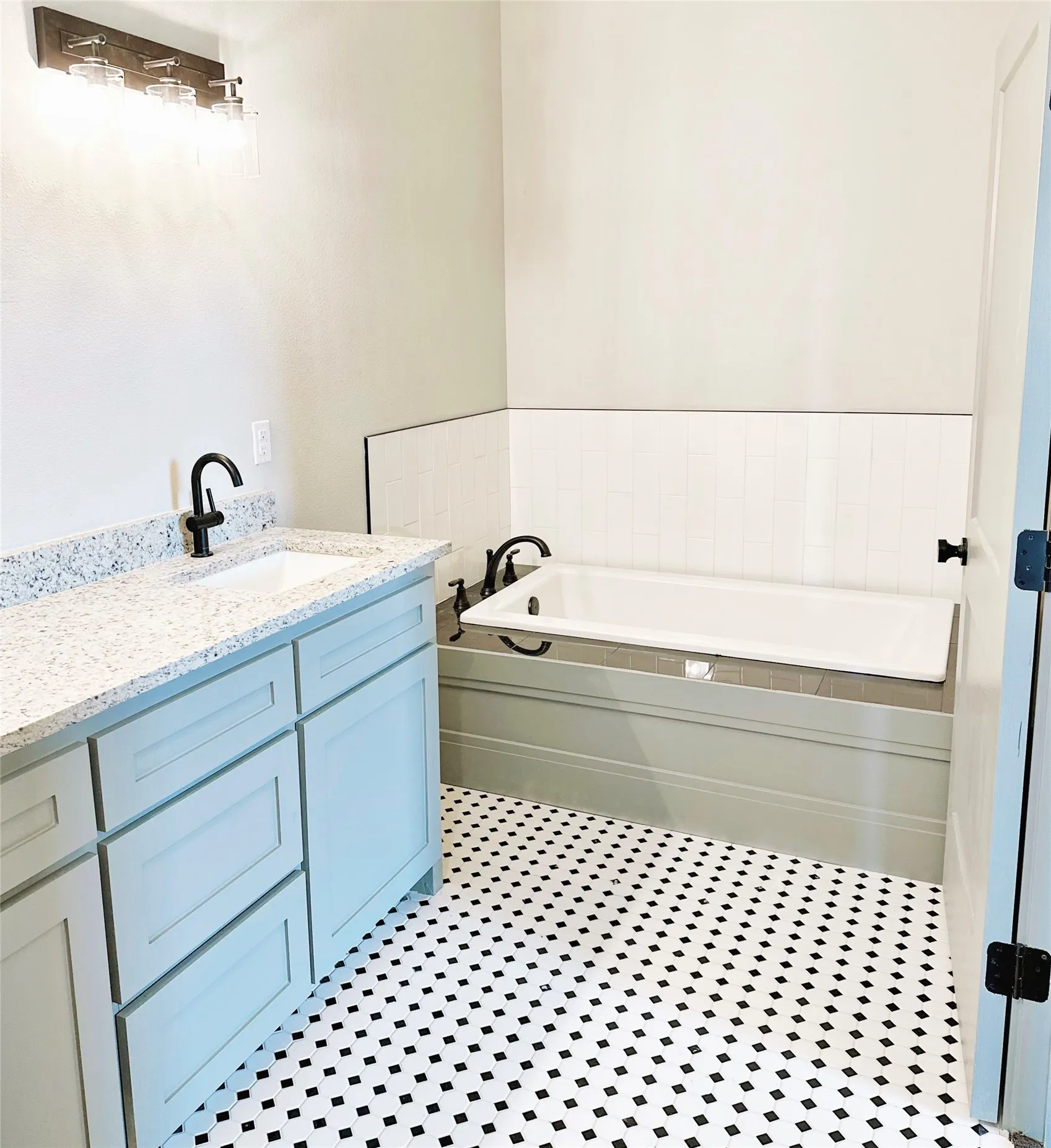 Bathroom featuring black and white mosaic floor tile, a light gray vanity with a single sink and stone-finish countertop, a tub with a light gray panel, and matte black fixtures