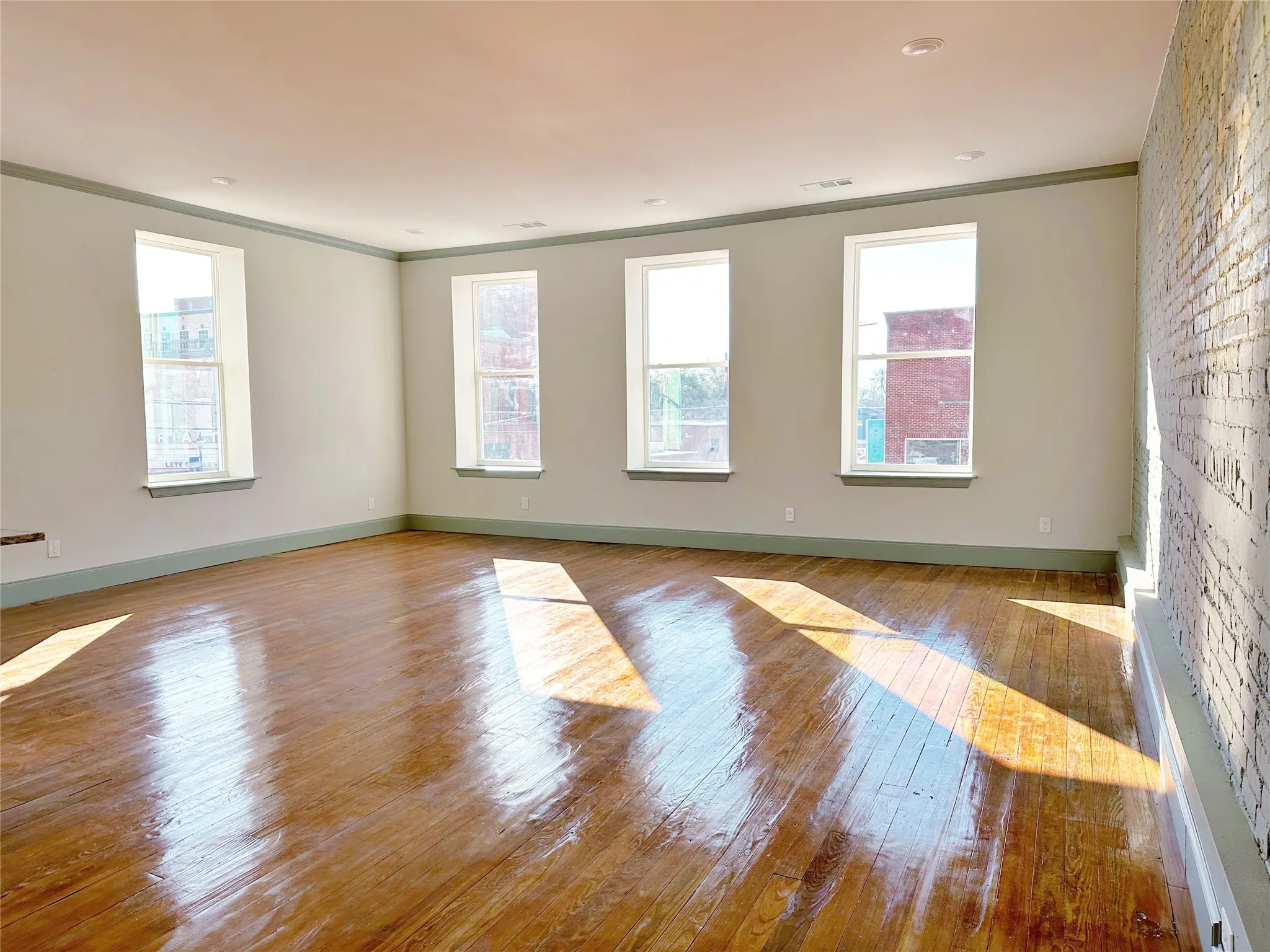 Spacious room featuring wood-finish flooring, a painted brick accent wall, four large windows, recessed ceiling lighting, and crown molding
