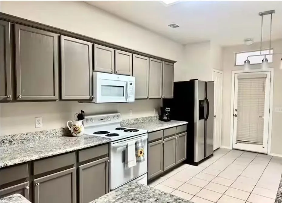 Kitchen featuring gray shaker-style cabinetry with crown molding, light-toned granite countertops, white tile flooring, and a stainless steel refrigerator