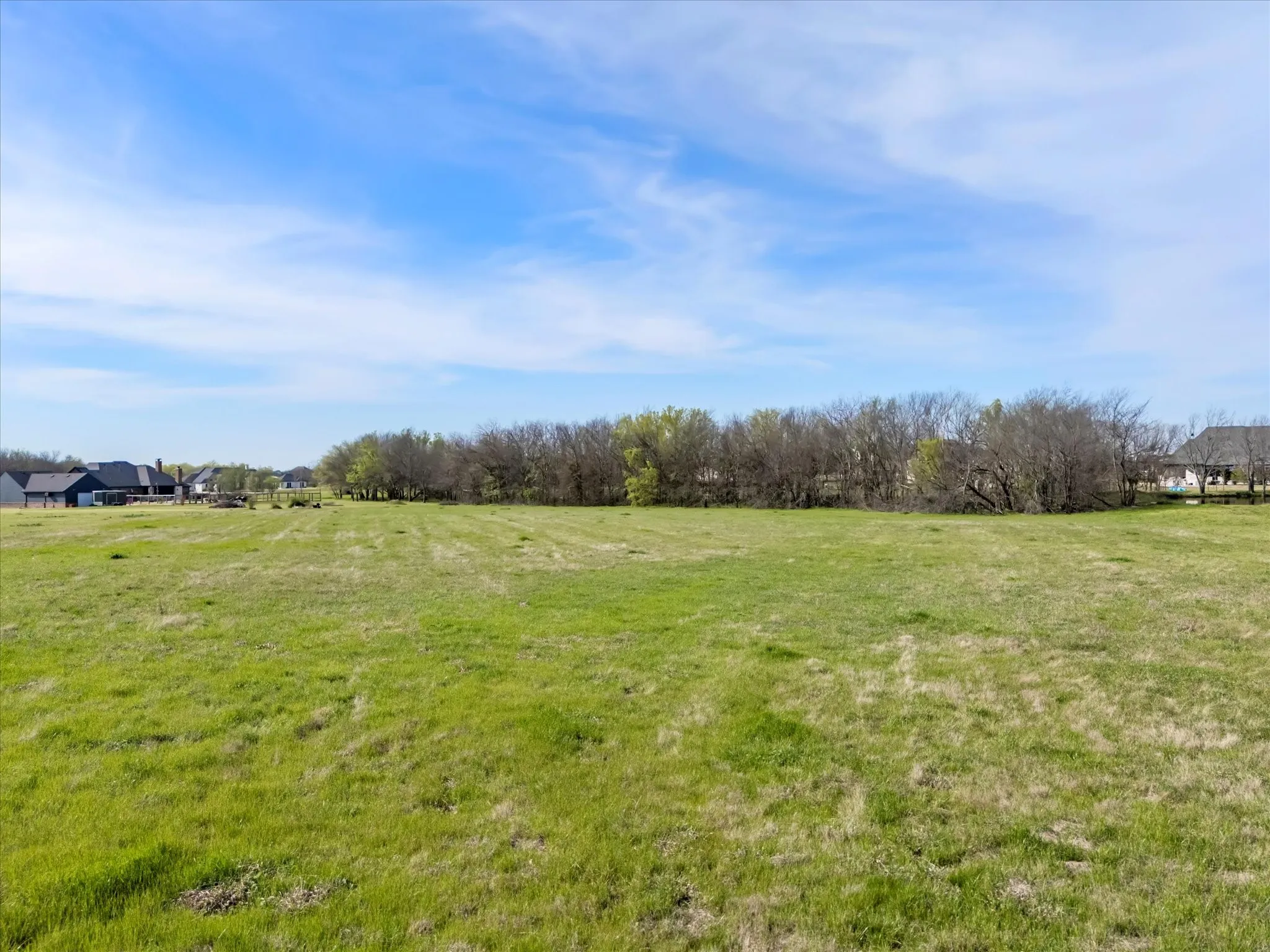 Expansive green field with a treeline border and distant residential structures under a clear sky