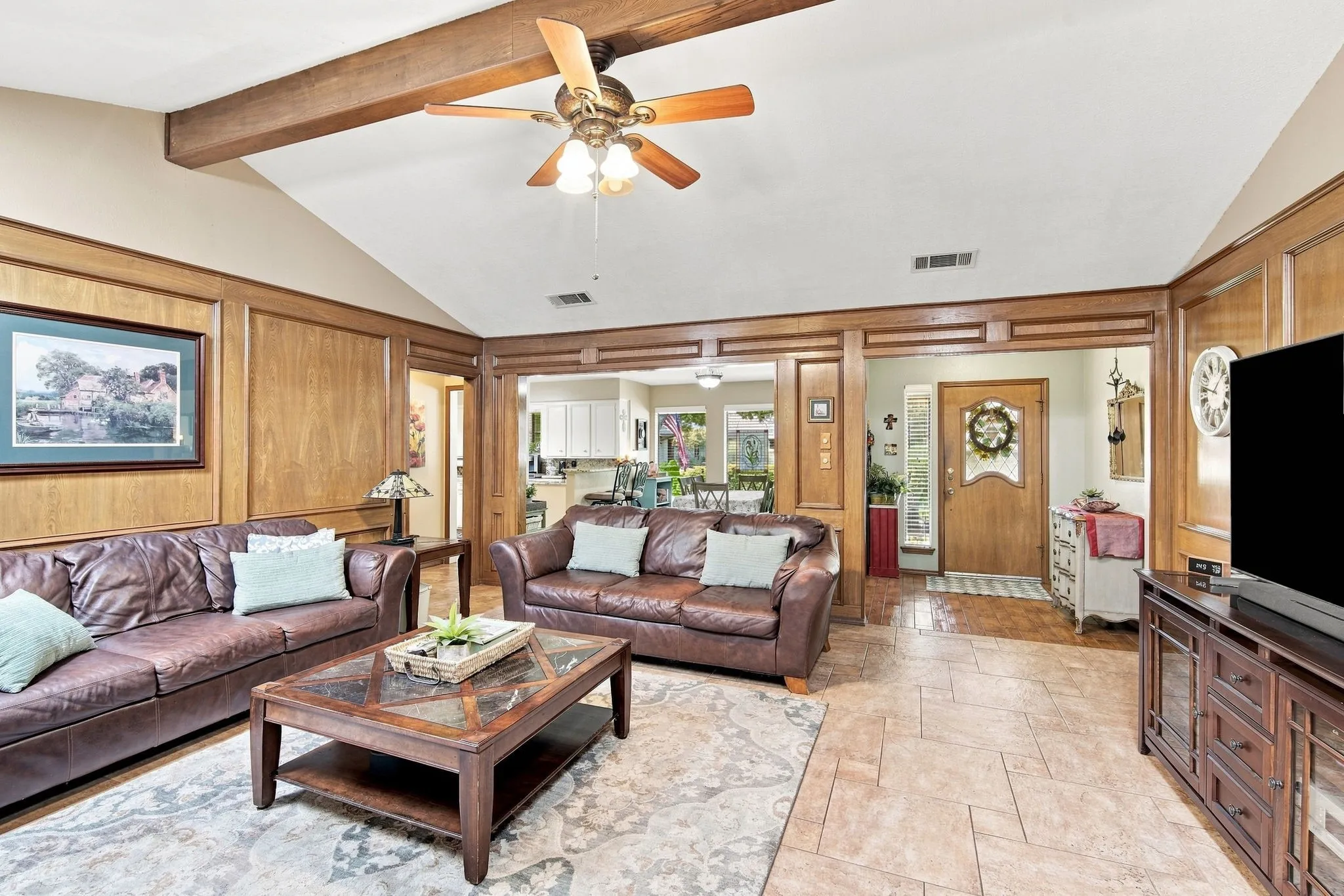 Spacious great room showcasing exposed wood beam ceiling with ceiling fan, dual leather sofas, wood-finish flooring, and extensive built-in cabinetry