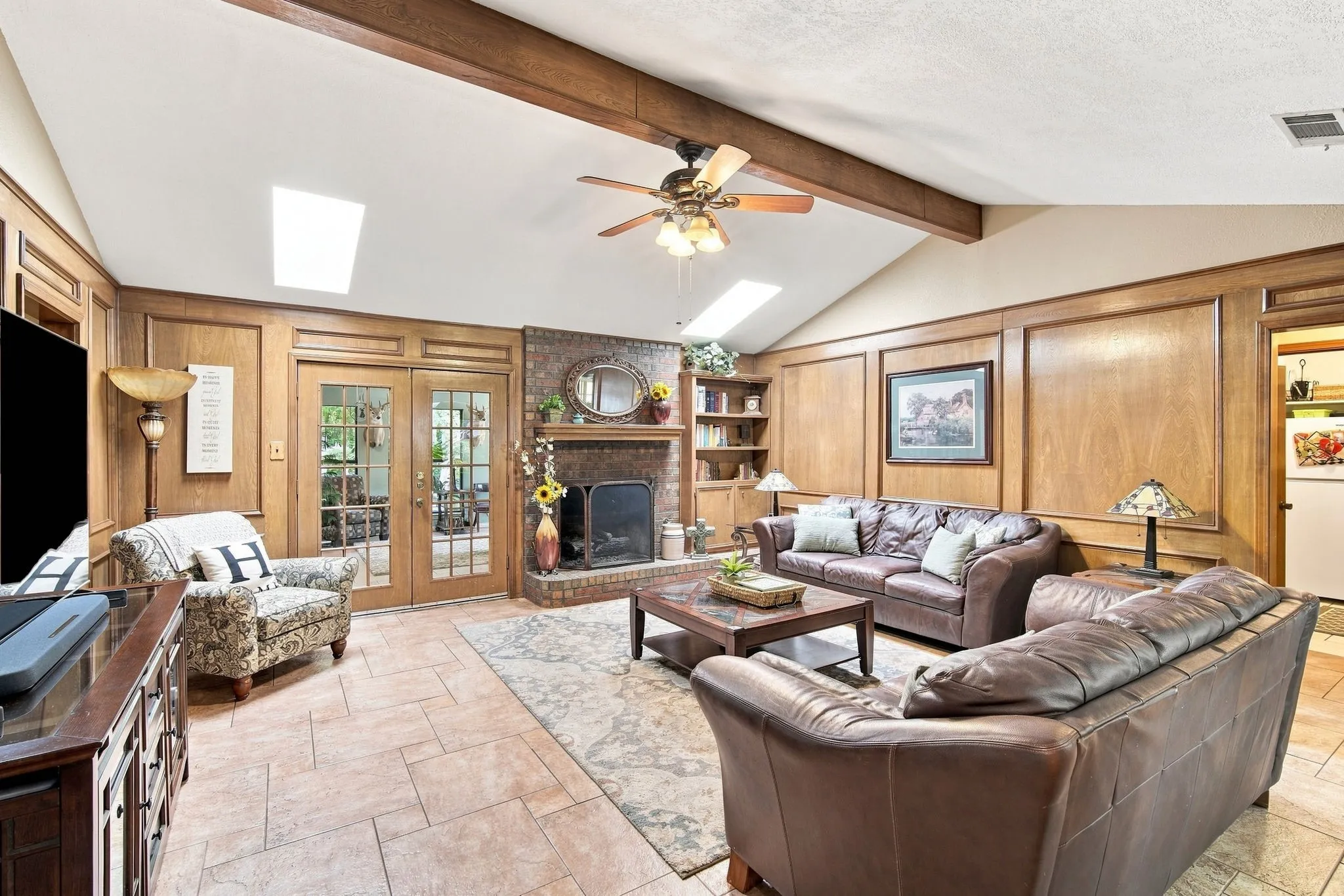 Vaulted ceiling featuring exposed wood beams and skylights, brick fireplace with built-in shelving, wood paneled walls, and French doors