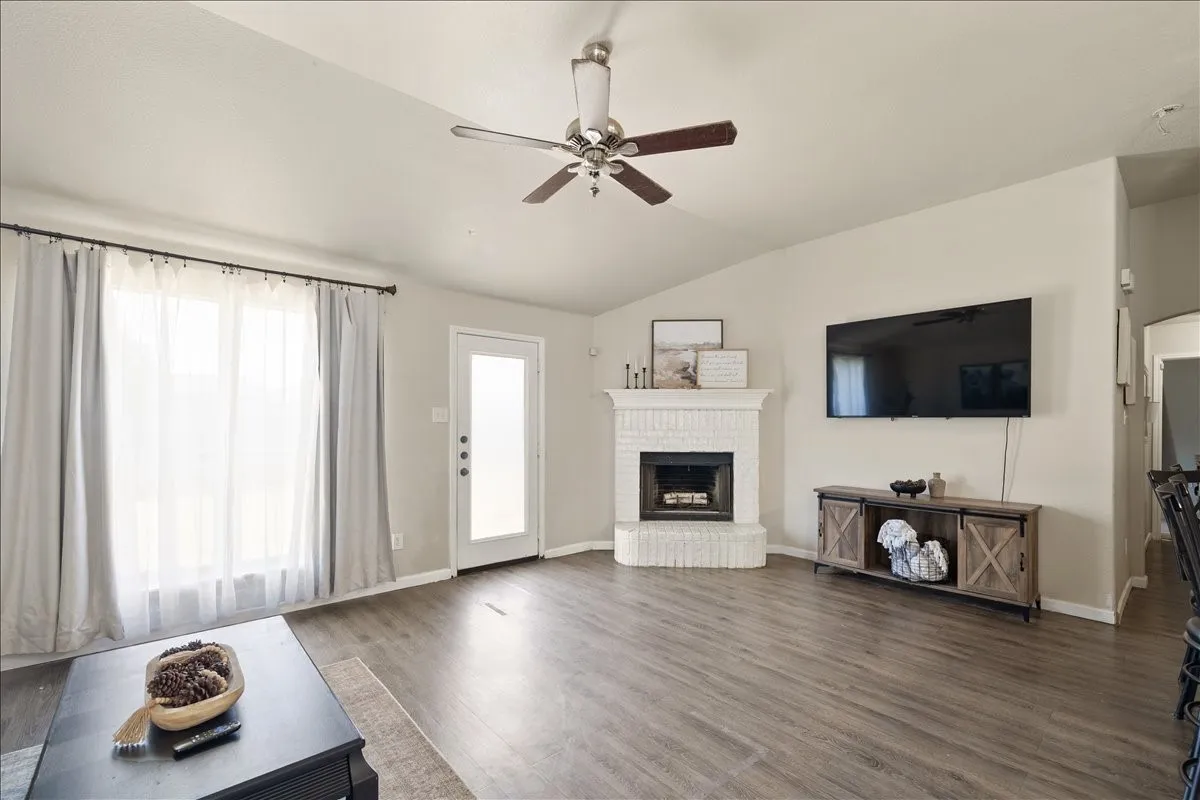 Spacious living area featuring vaulted ceilings, wood-finish flooring, a white brick fireplace, and a ceiling fan with light fixture