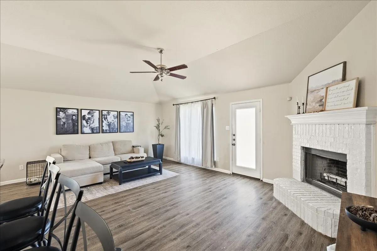 Vaulted ceiling living area featuring wood-finish flooring, a painted brick fireplace with hearth, and a ceiling fan with light fixture