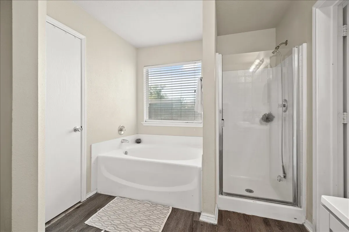 Bathroom featuring a white soaking tub with polished chrome fixtures, a separate shower enclosure with a glass door, a window with blinds, and wood-finish flooring