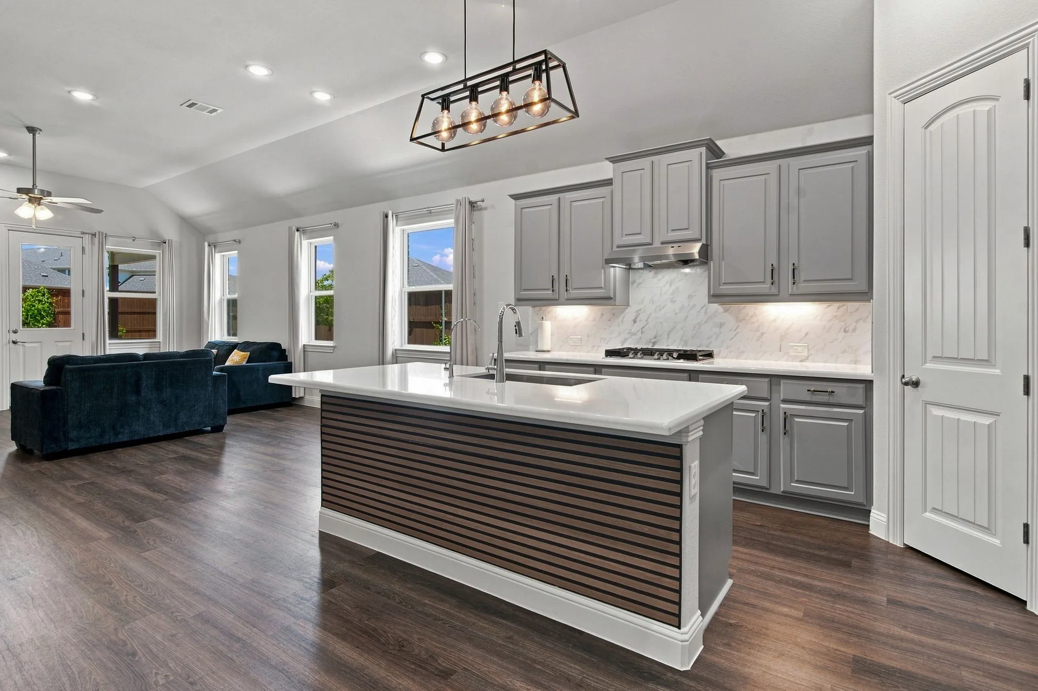Open-concept kitchen and living area featuring wood-finish flooring, vaulted ceilings, a central kitchen island with a slatted wood-look accent, gray cabinetry, and white countertops with a tiled backsplash