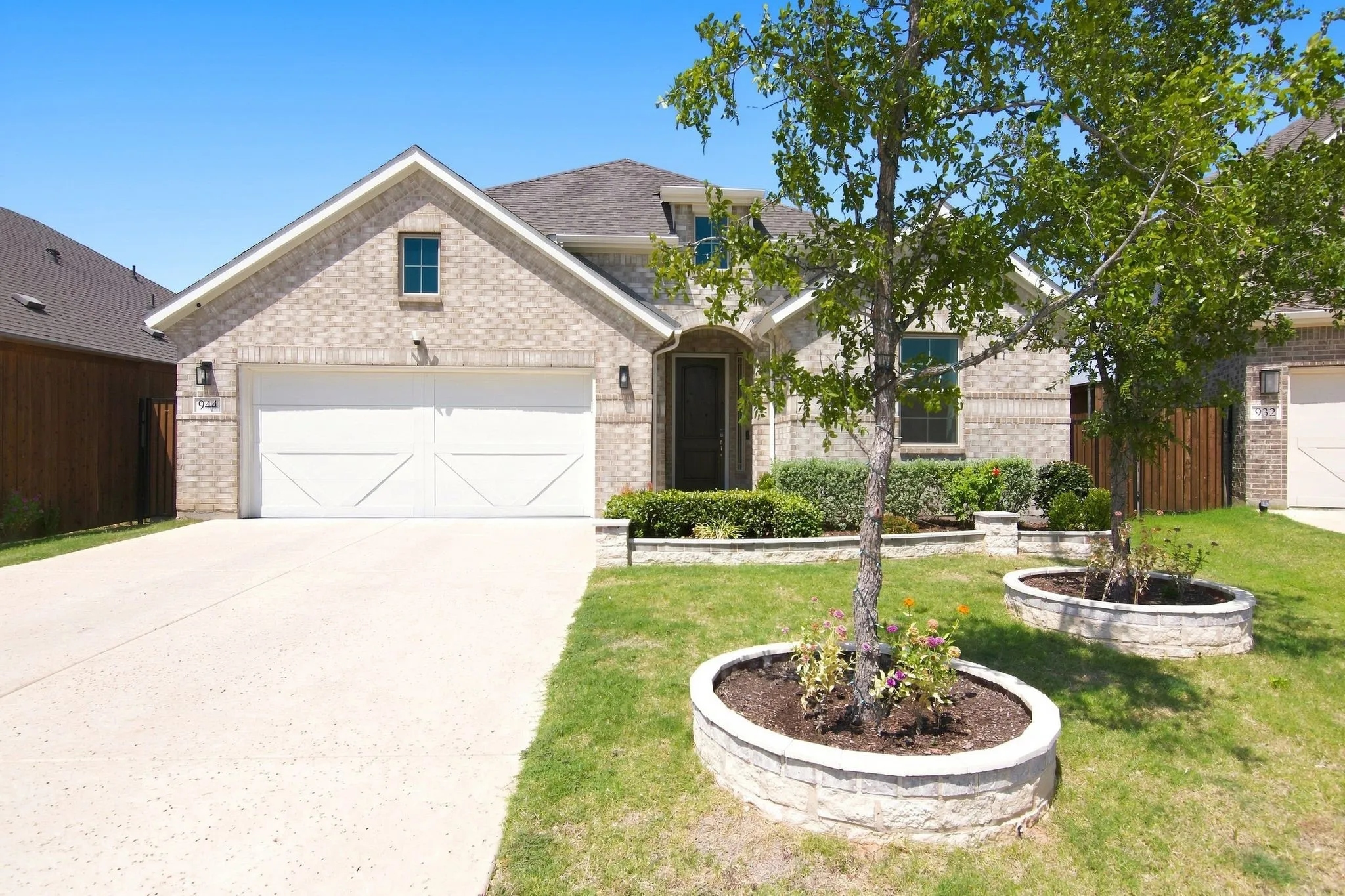 Brick facade residence featuring a two-car garage with white paneled doors, an arched entry, a concrete driveway, and landscaped planting beds with stone retaining walls
