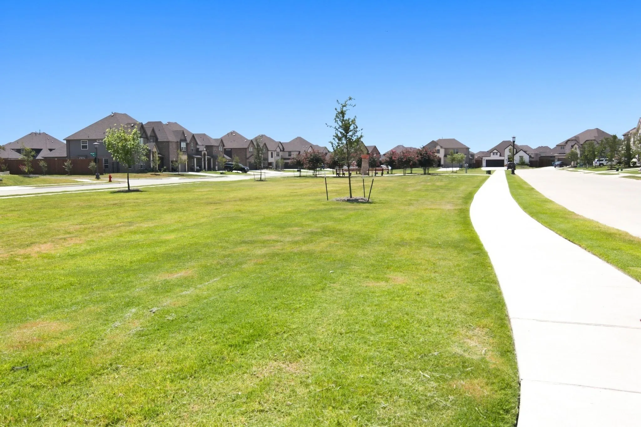 Expansive green space featuring a paved walking path, scattered young trees, and a backdrop of residential homes