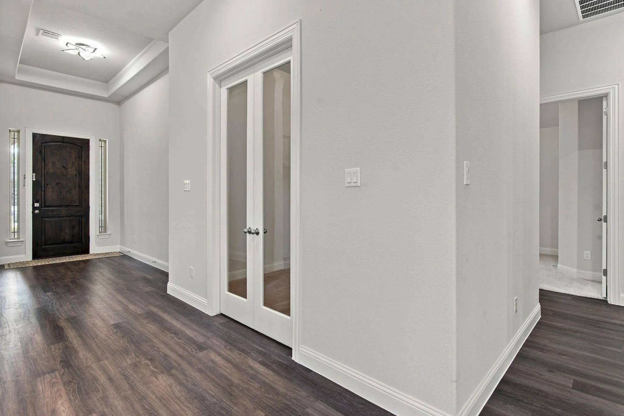 Entryway featuring wood-finish flooring, a dark wood front door with sidelight windows, a tray ceiling with an attached light fixture, and white double French doors with frosted glass panels