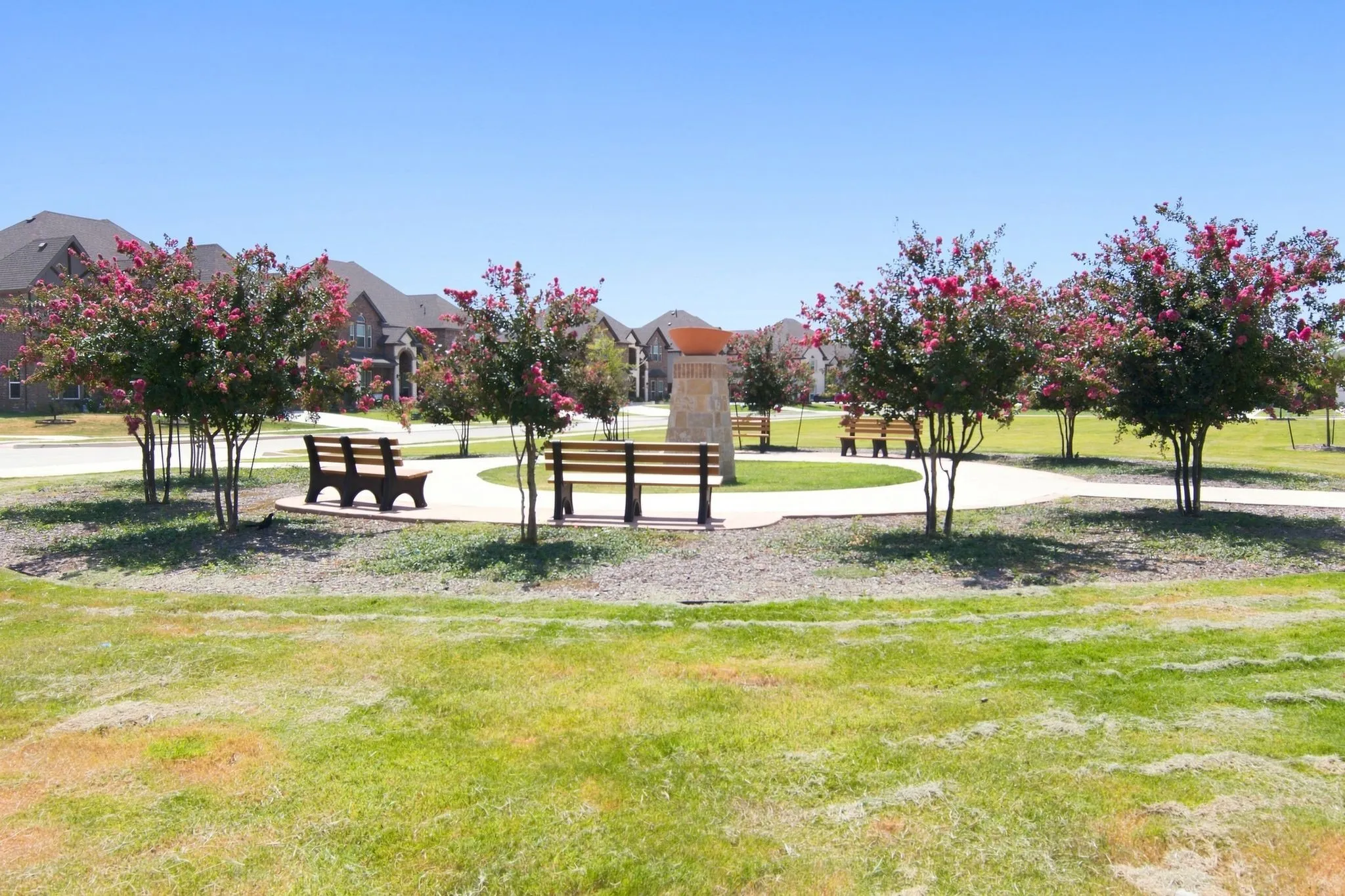 Community green space featuring a circular walking path, multiple park benches, mature flowering trees, and a central stone monument