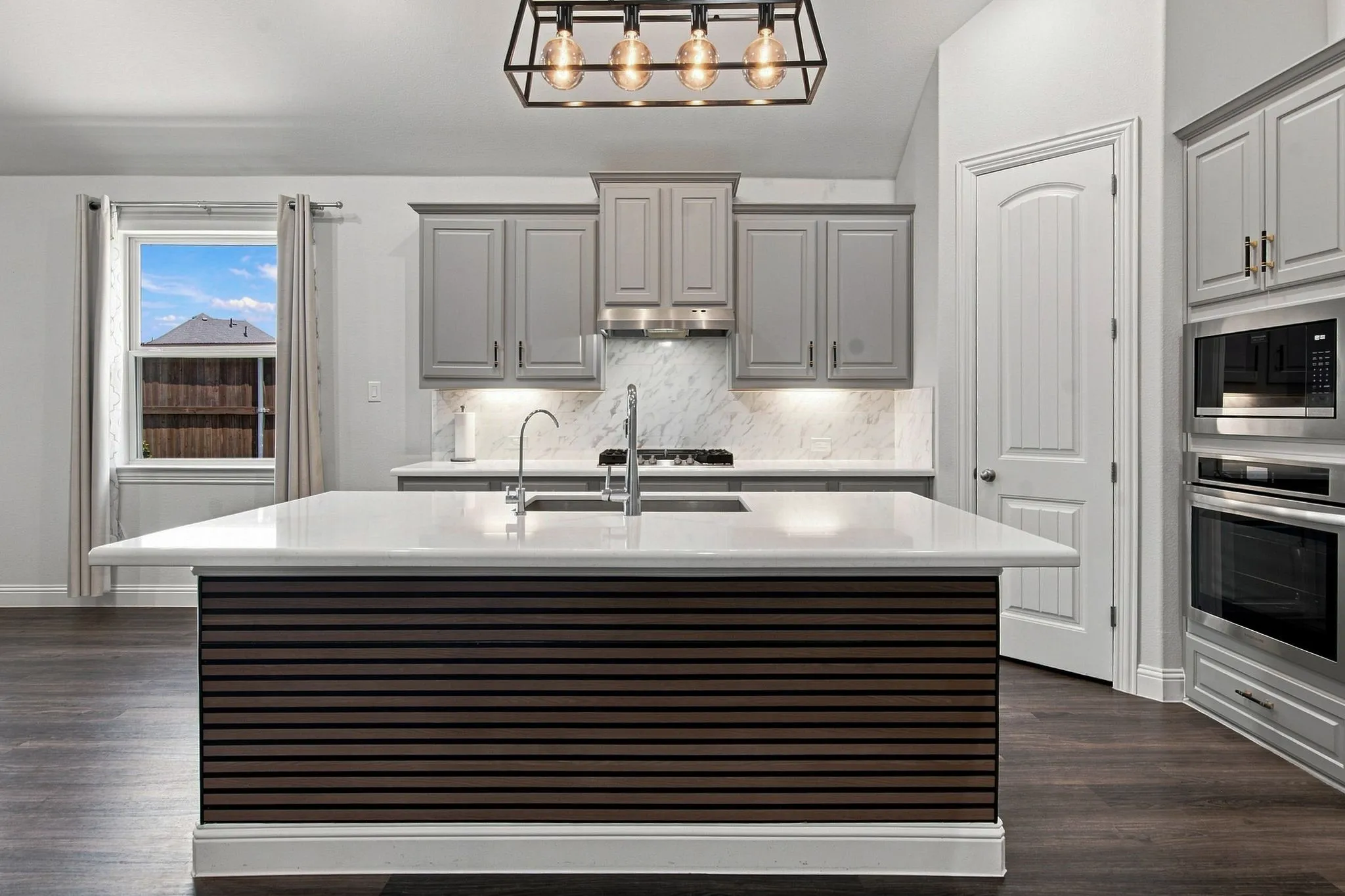 Spacious kitchen featuring a large island with a slatted wood-finish base and light-toned countertop, gray cabinetry, built-in stainless steel appliances, a range hood, and wood-finish flooring