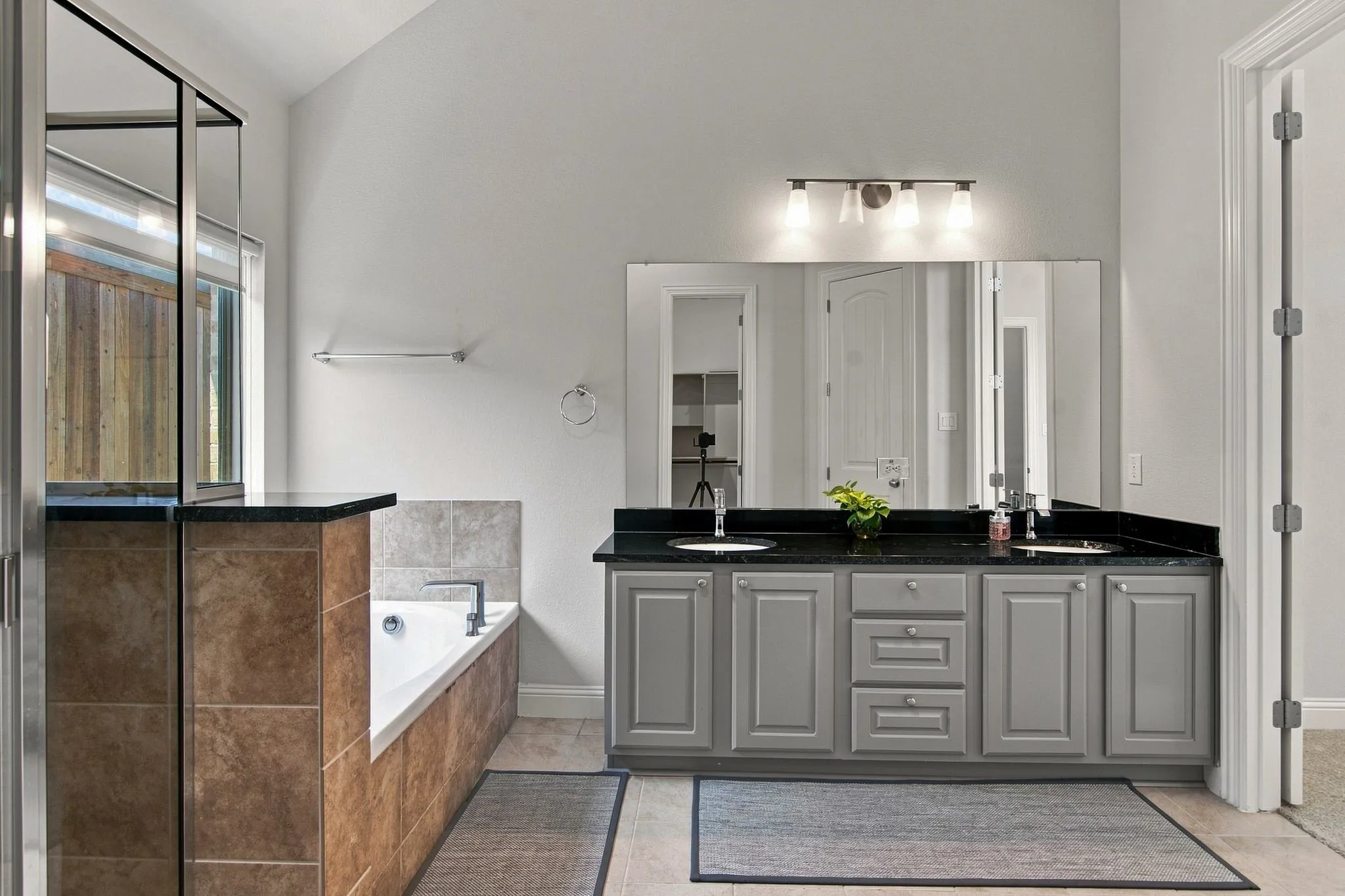 Spa-inspired primary bathroom featuring a dual-sink vanity with gray cabinetry, black countertops, and polished hardware, complemented by a frameless glass-enclosed shower with black accents, large mirror with integrated lighting, and neutral tile flooring