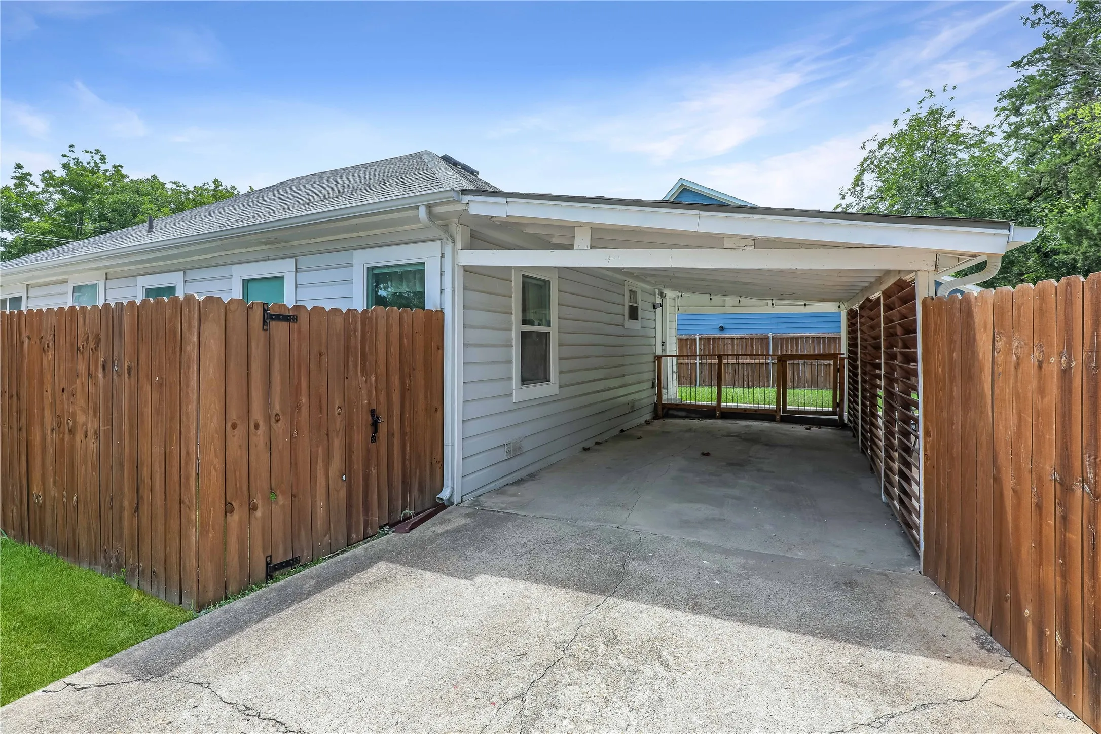 Carport with concrete driveway, featuring white siding, a wood fence, and a gate leading to a rear yard