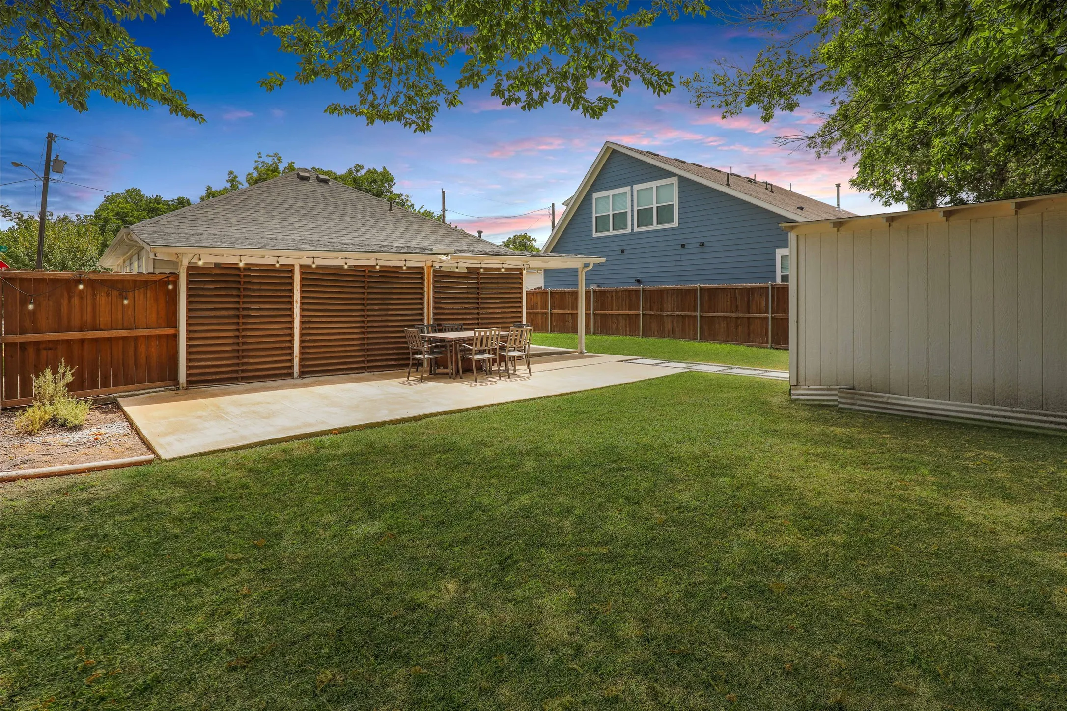 Expansive rear yard featuring a concrete patio, covered pergola with slatted wood walls, and a separate storage shed