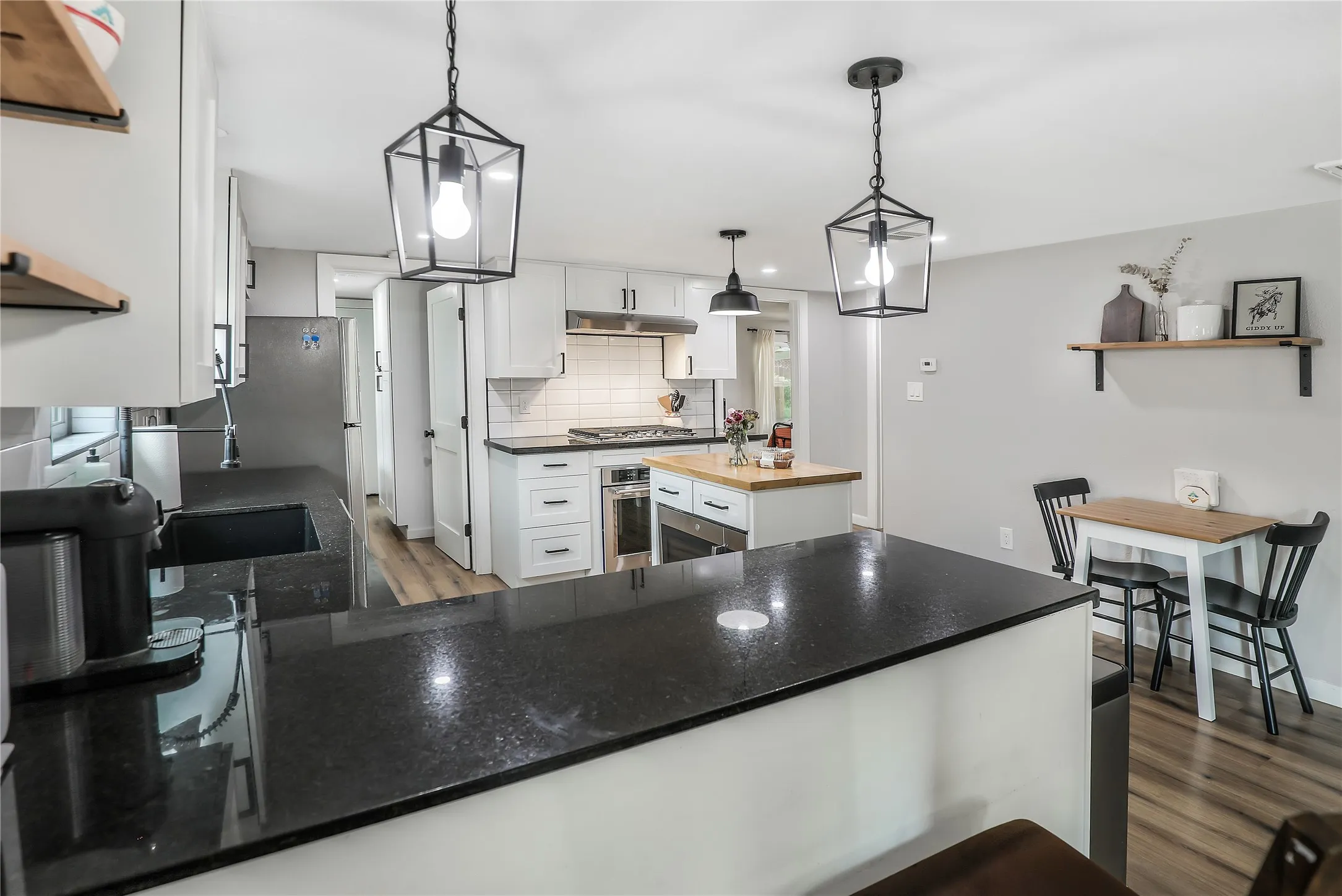 Modern kitchen featuring black countertops, white shaker cabinetry, a central island with a wood-finish top, wood-finish flooring, and pendant lighting