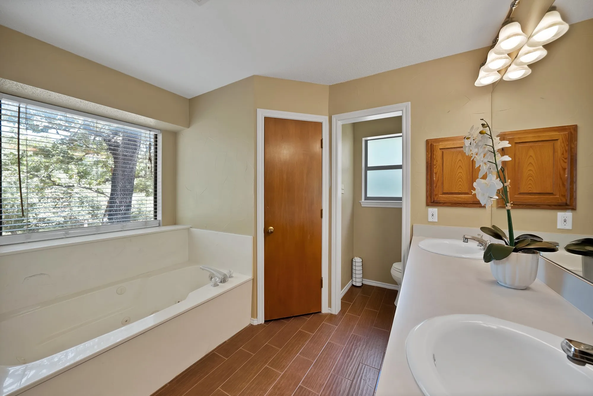 Soaking tub with surrounding window, dual vanity with undermount sinks, wood-finish flooring, and natural wood cabinetry