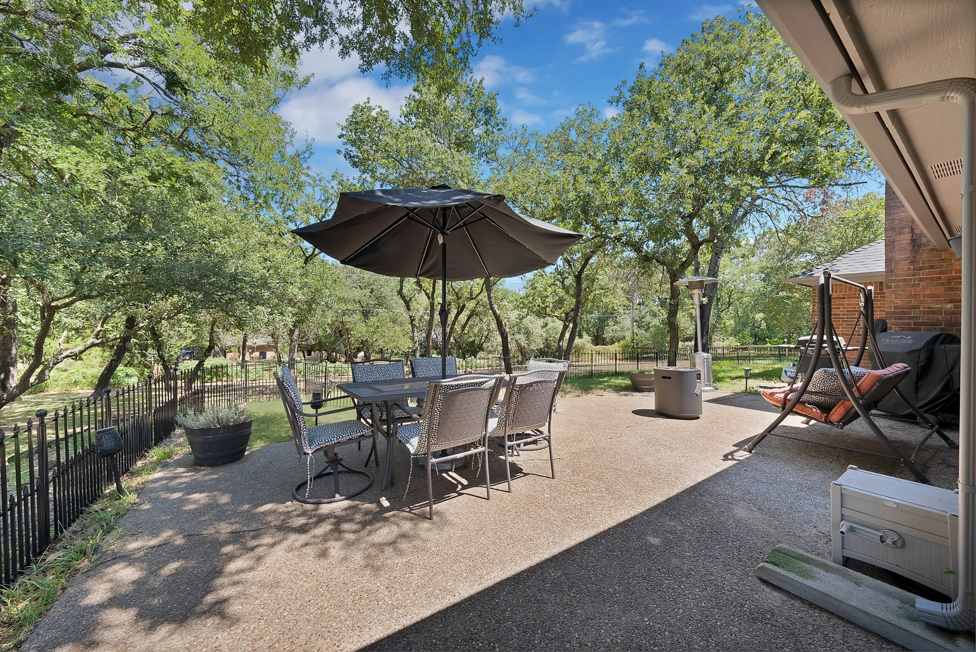 Expansive patio with exposed aggregate concrete flooring, bordered by a black metal fence and mature trees