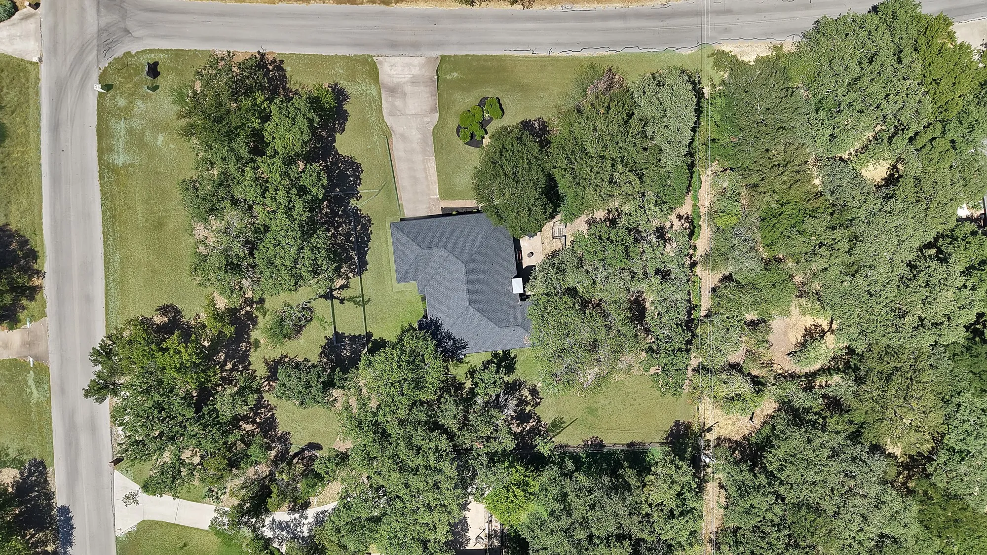 Corner lot property featuring a dark-toned shingle roof, concrete driveway, and extensive mature trees