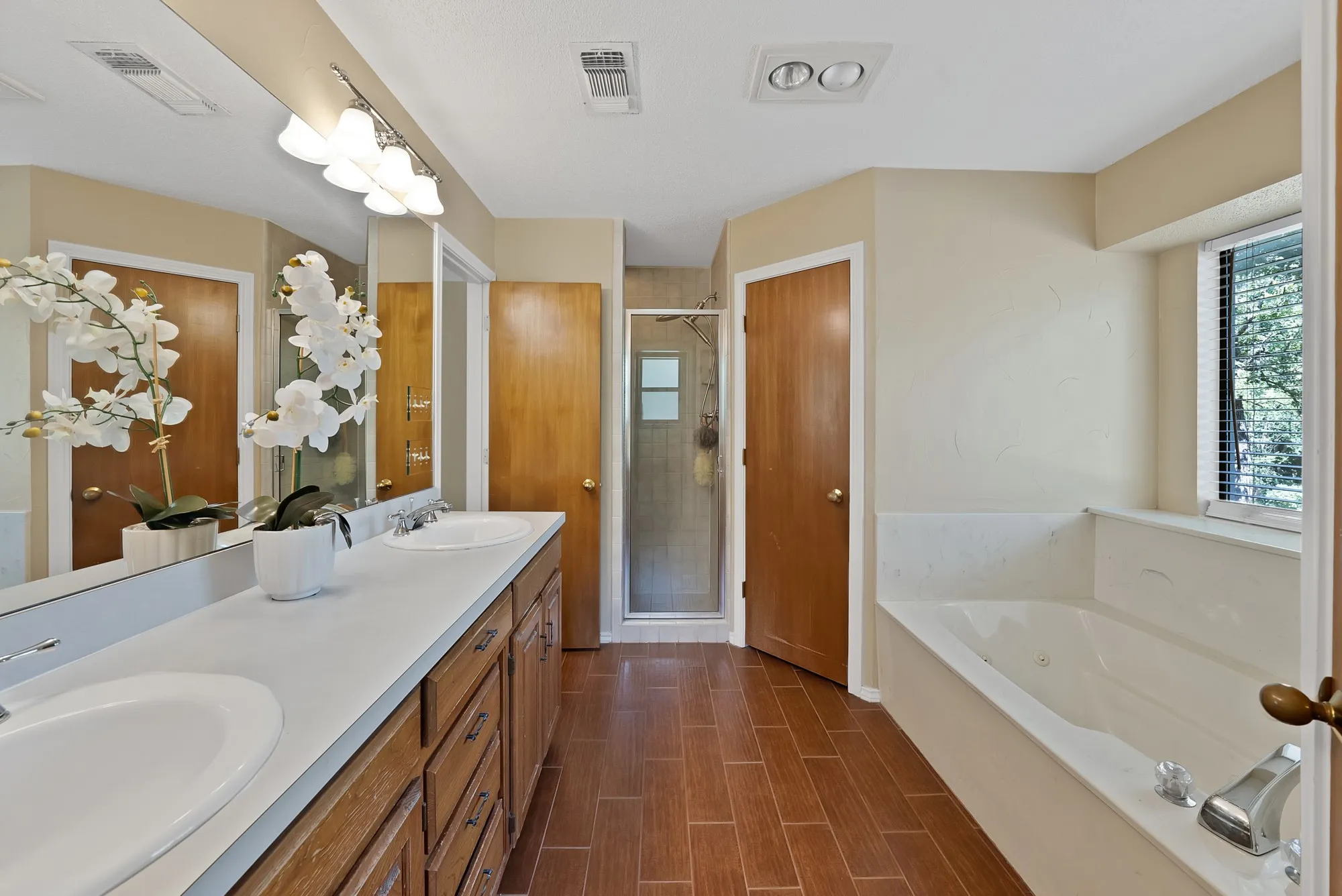 Dual vanity with integrated sinks, full-width mirror, wood-finish flooring, built-in soaking tub, and enclosed shower