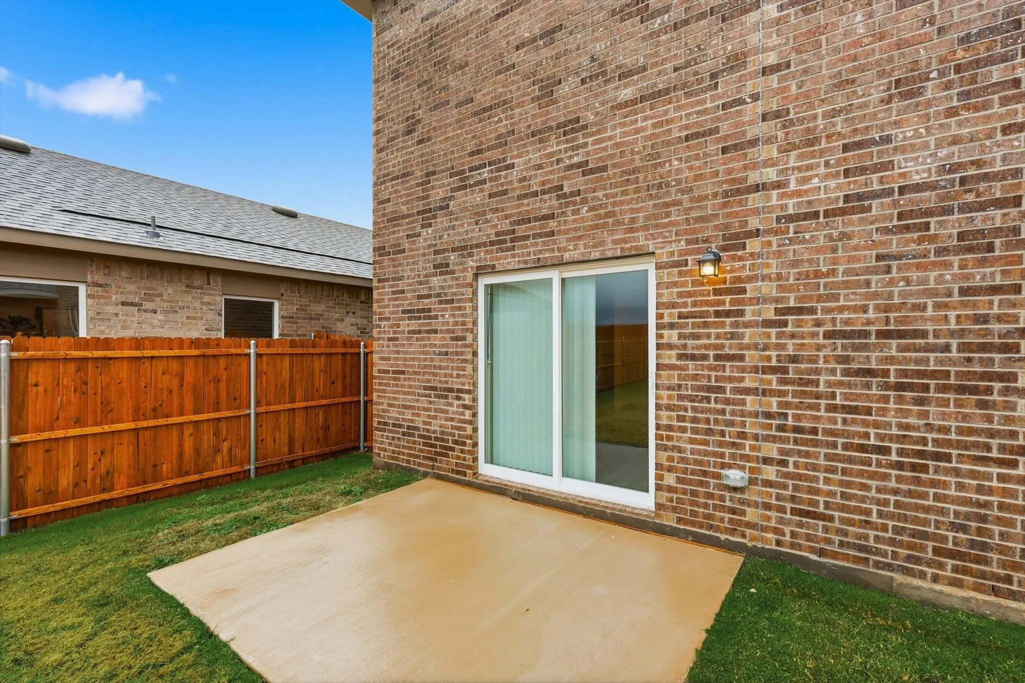 Brick exterior featuring a sliding glass patio door, concrete patio, a wall-mounted light fixture, and a wood privacy fence