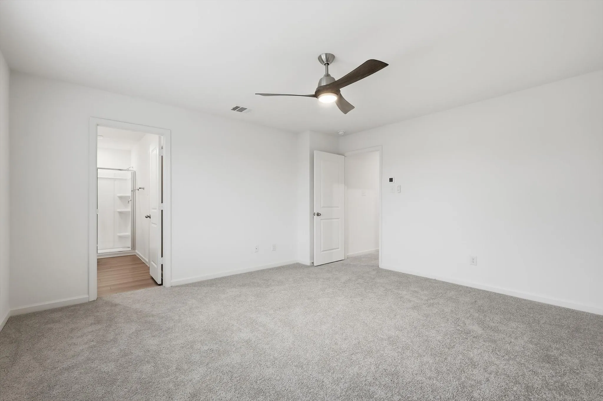 Spacious room featuring light gray carpeting, crisp white walls, a contemporary ceiling fan with integrated lighting, and a doorway revealing a bathroom with wood-finish flooring and a shower enclosure