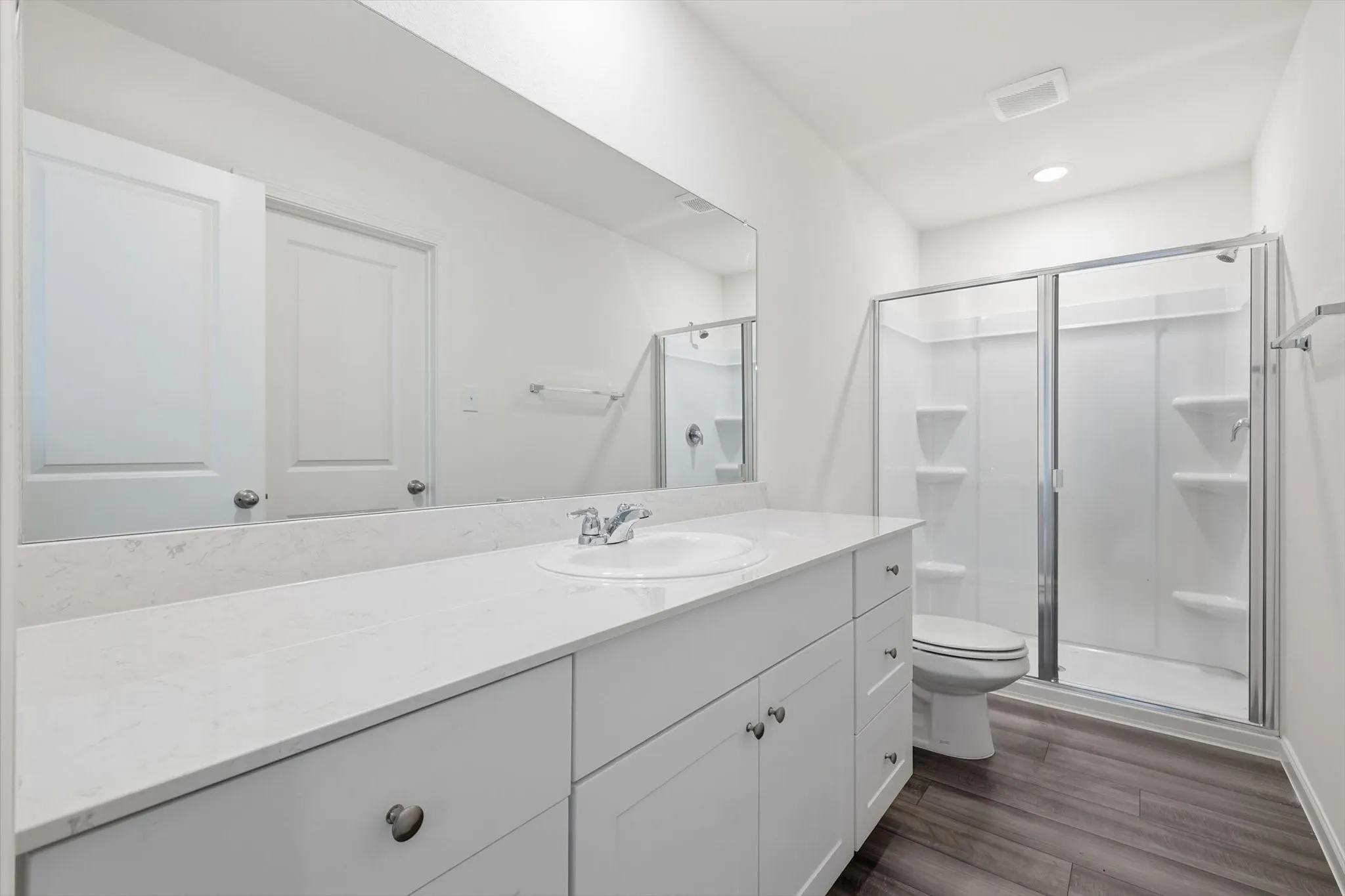 Bathroom featuring an expansive vanity with white cabinetry and a light-toned countertop, a large wall-mounted mirror, a glass-enclosed shower with a white surround, and wood-finish flooring